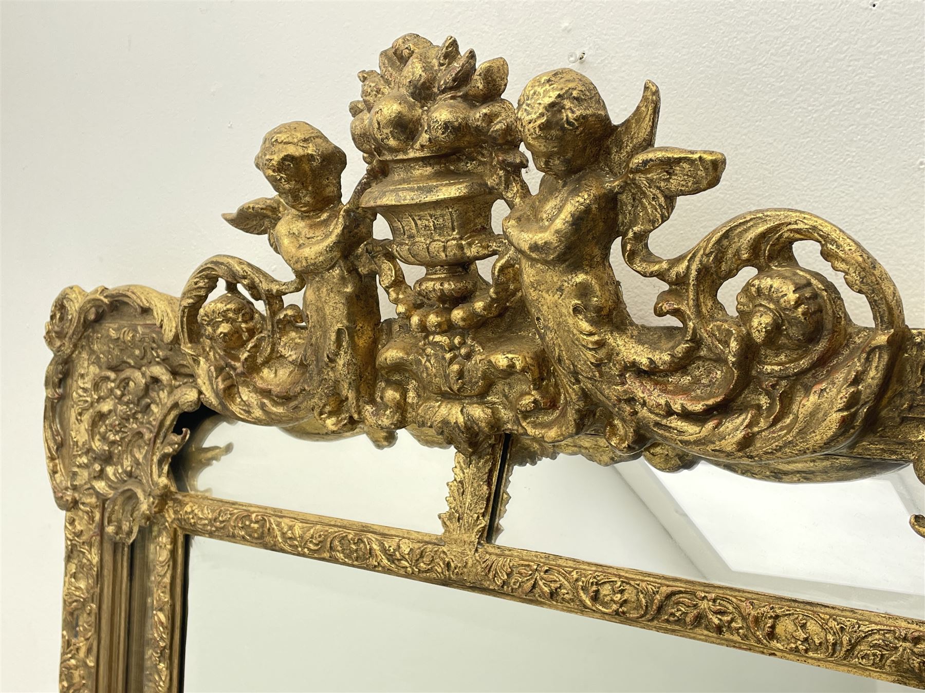 Large Victorian style gilt cushion framed mirror, putti holding urn with scrolled foliage pediment above central rectangular bevelled plate, the frame decorated with moulded foliate, each corner set with ornate cartouche
