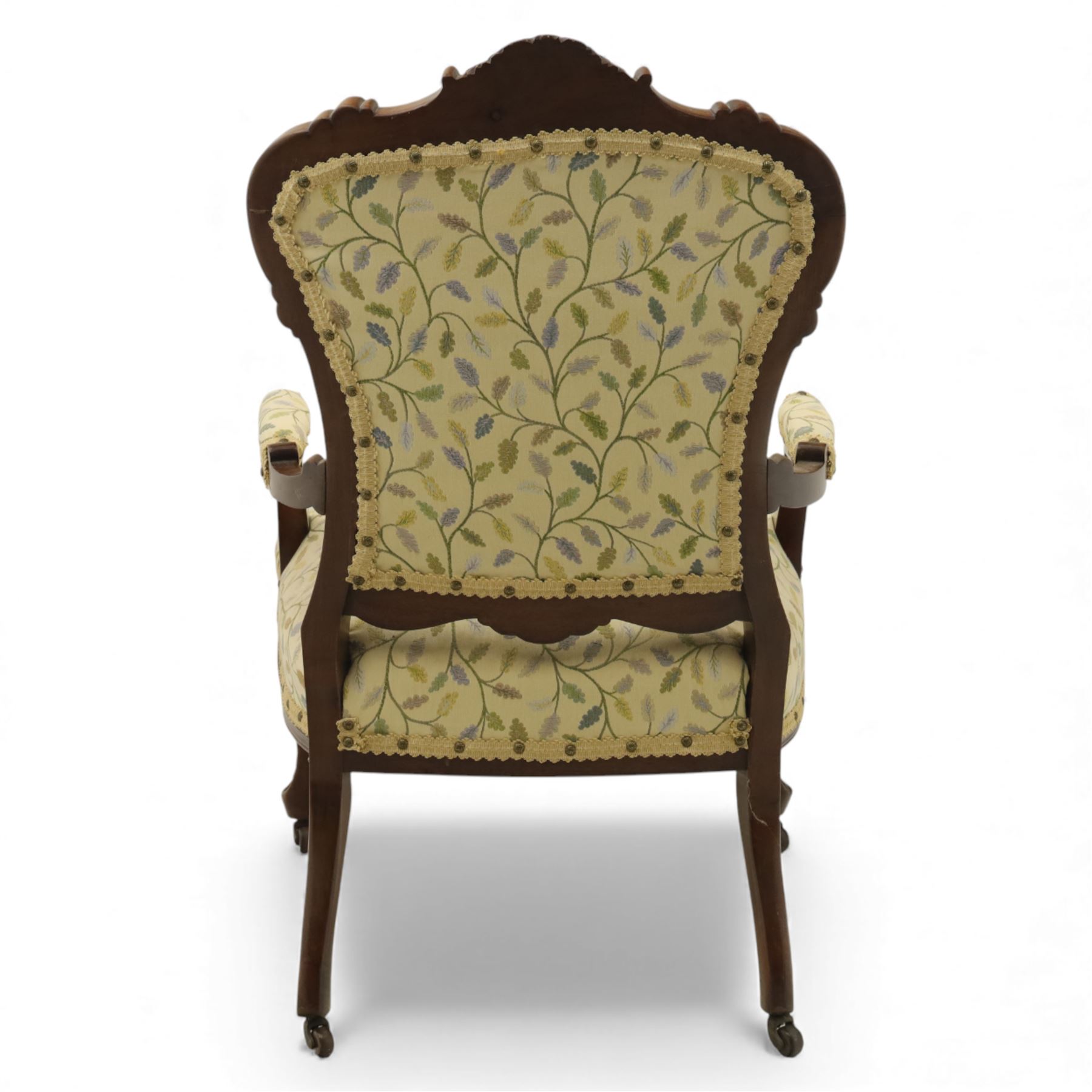 Victorian mahogany framed upholstered armchair, shaped and scroll carved cresting rail above a padded upholstered back with applied braid trim, swept open arms with carved terminals and upholstered arm pads, over a serpentine fronted deep stuffed seat, raised on cabriole forelegs terminating in carved claw feet