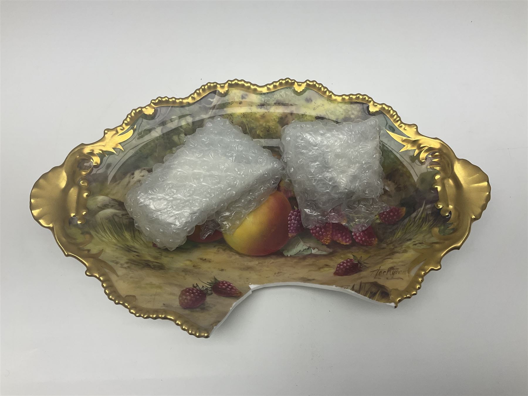 Early 20th century Royal Worcester dish decorated by Thomas Lockyer, oval form with gilt shaped rim and twin shell handles, hand painted with a still life of fruit upon a mossy ground, signed T Lockyer, with puce printed mark beneath and date code for 1922, for restoration, W31cm