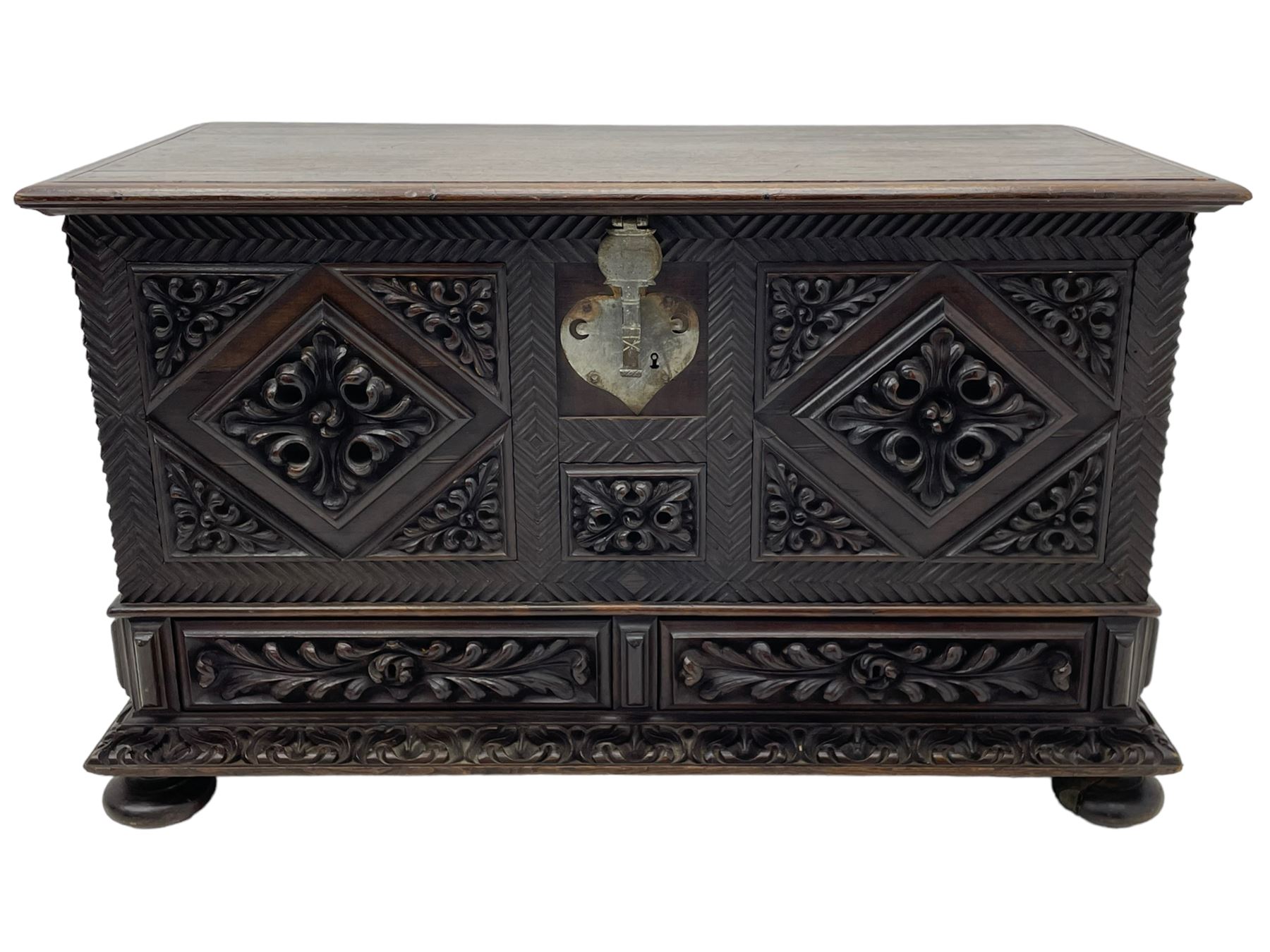 Early 20th century carved hardwood kist or mule chest of small proportions, moulded rectangular hinged lid over foliage carved geometric lozenge panels, the frame carved with chevron banding, fitted with two drawers, the drawer fronts carved with extending leaf motifs over a foliage carved lower moulding, on turned feet