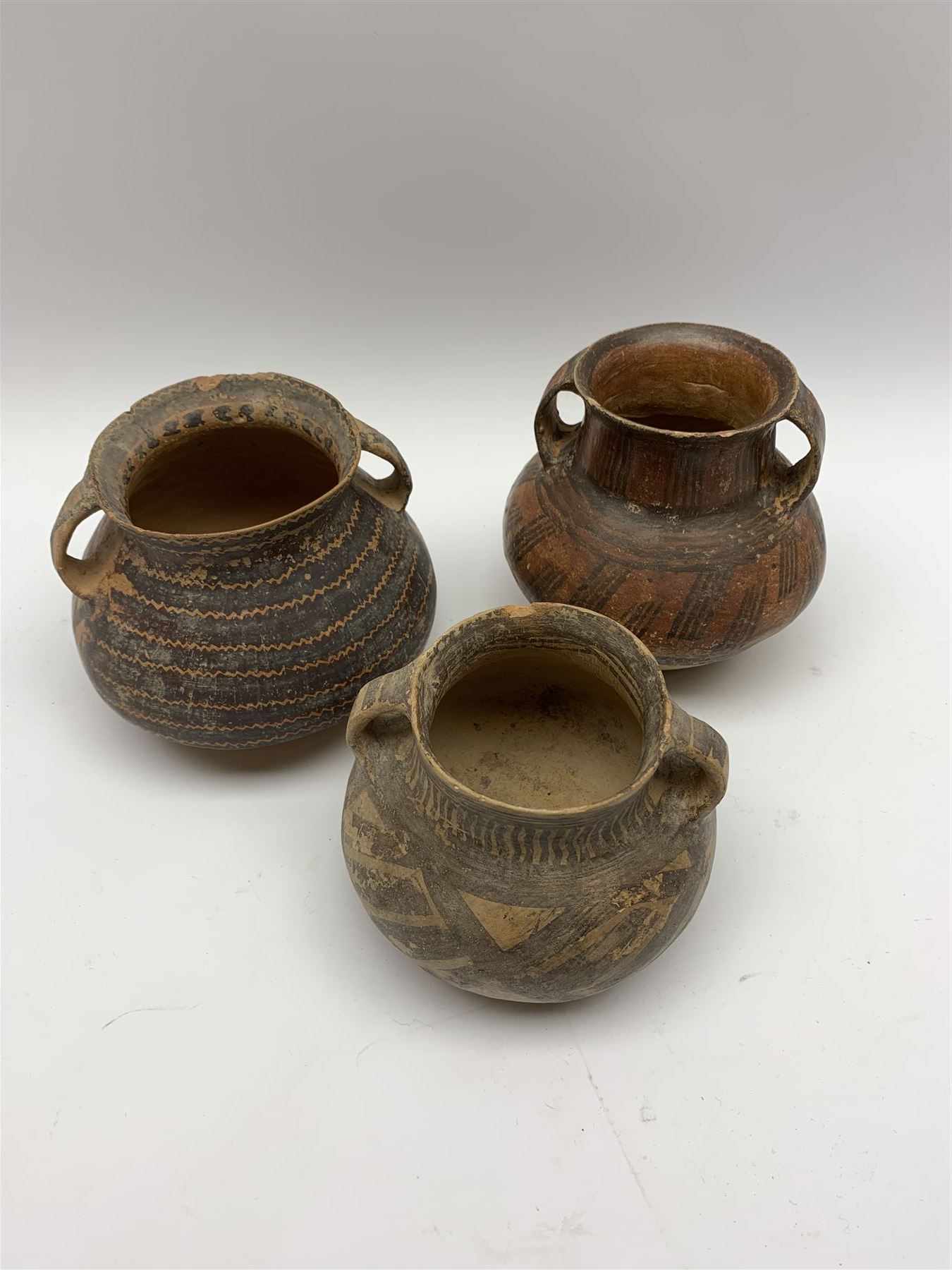 Three Chinese Neolithic pottery vessels, each with twin loop handles to the shoulder and painted geometric banded decoration to the body, all bearing labels for Eskenazi London, tallest H14cm