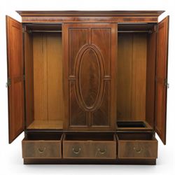 Edwardian inlaid mahogany triple wardrobe, featuring two oval mirror doors with bevelled plates, enclosing central figured veneered panel with mouled detail, above three base drawers