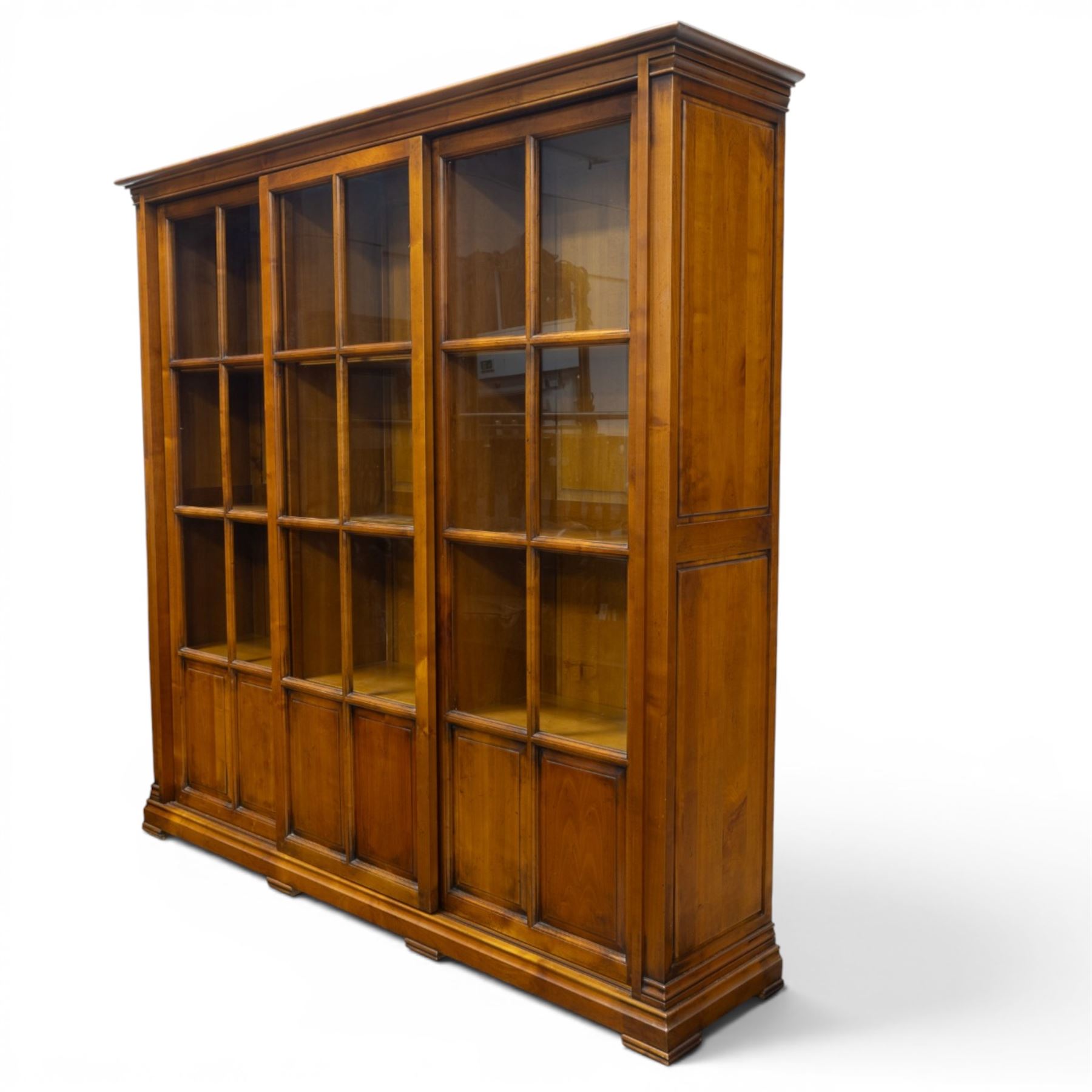 Wesley Barrel - large cherry wood bookcase, with projecting cornice and fielded panelled sides, with three sections comprising three fixed shelves, sliding glazed and panelled doors, moulded plinth base