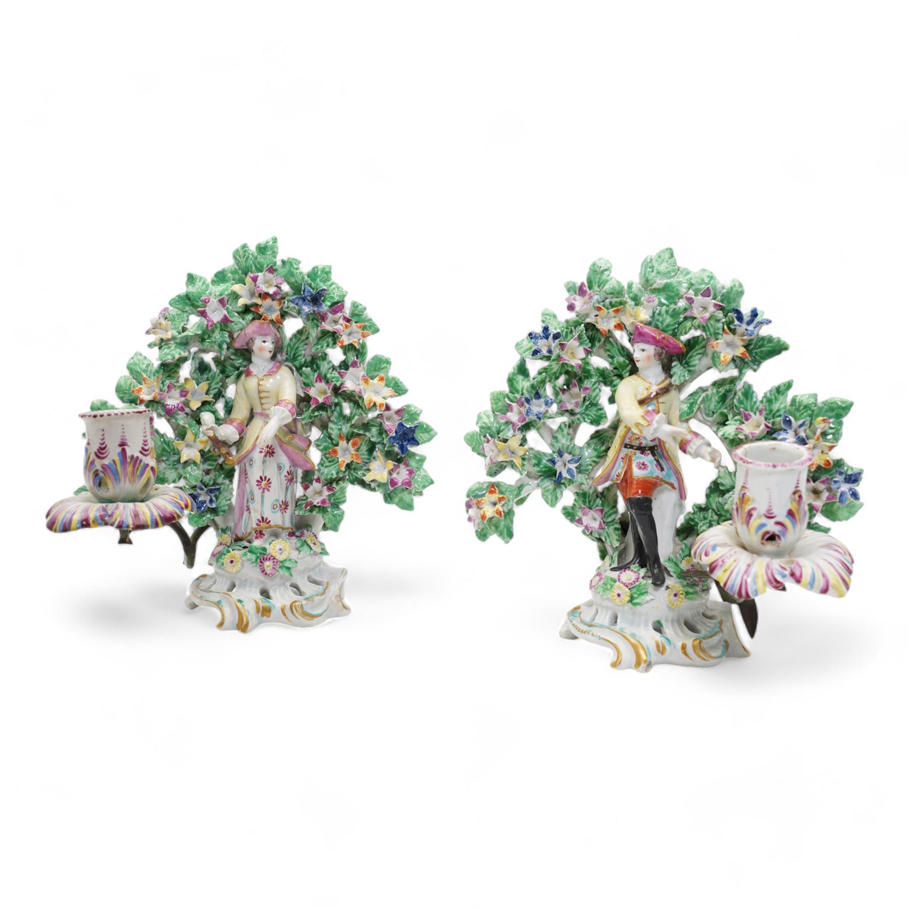 Pair of Bow porcelain 'New Dancers' candlesticks, standing before flowering bocages, and wearing brightly coloured flowered clothes, each supporting to left and right a tôle peinte branch terminating in a leaf moulded nozzle, on scrolling footed bases, red anchor and blue/red dagger marks beneath, H19cm x W18cm (2)