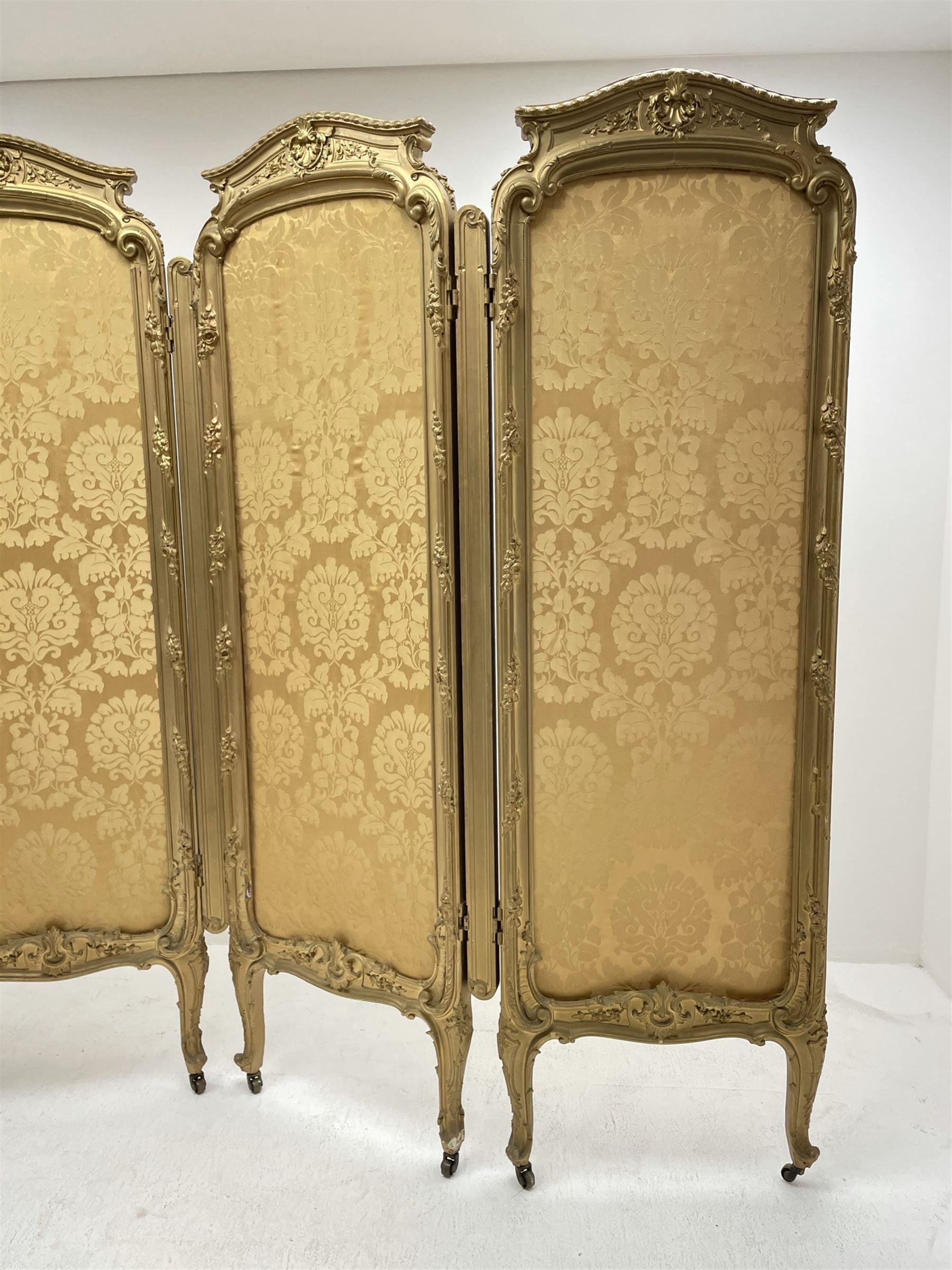 Late 19th/early 20th century French Rococo style gilt wood and gesso dressing screen, four double hinged panels, each with shaped cresting rail over shell and floral frieze, moulded frame with flower head and scroll decoration, acanthus leaf cabriole supports with castors, upholstered in gold Damask fabric