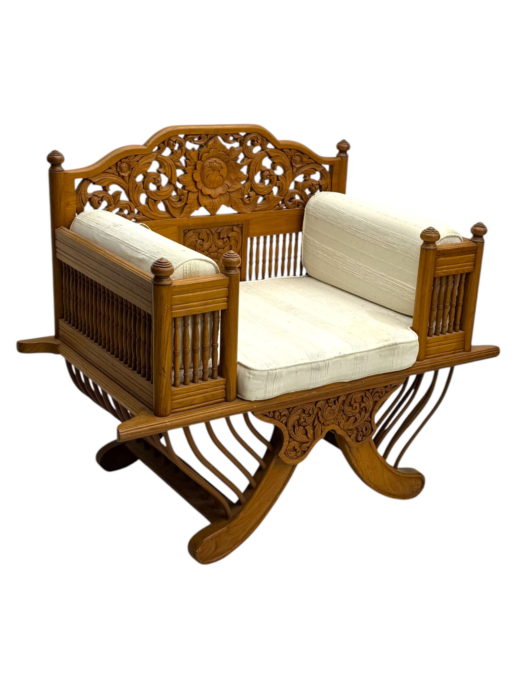 Early 20th century carved teak Howdah elephant chair, with pierced floral and foliate carved backrest, spindle gallery arm supports, cream upholstered cushions, splayed legs on a curved reeded base 