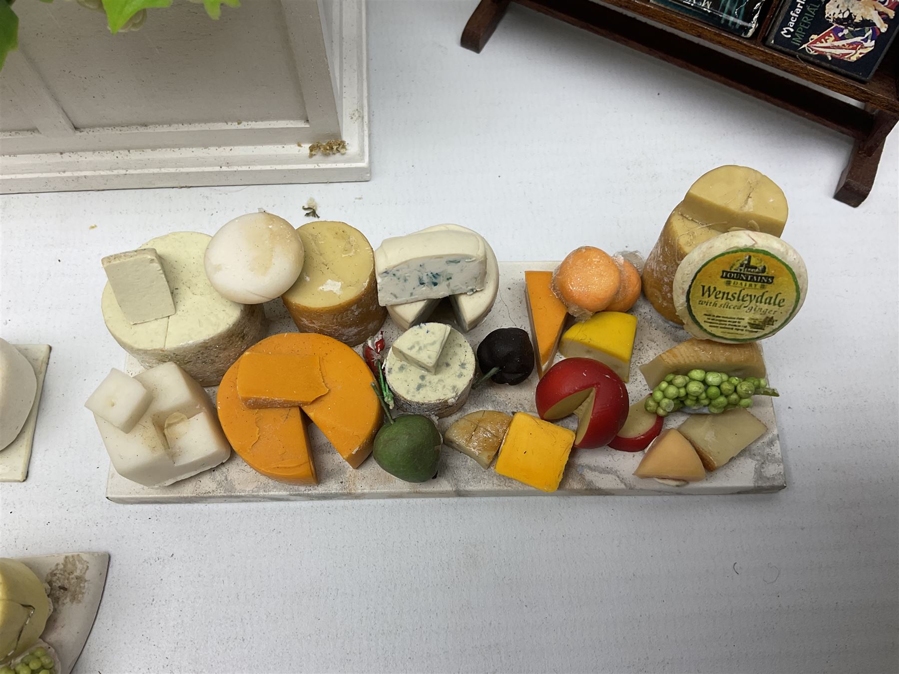 Collection of miniature dolls house grocery shop furniture and produce, comprising glazed corner counter with brass style cash register, shelved preserves unit, pies, cheese board and trays of meats, pair of white cheese counters, biscuit display with six tins, glazed preserves cabinet, pair of shop chairs and four further cheese boards complete with grapes and label flags