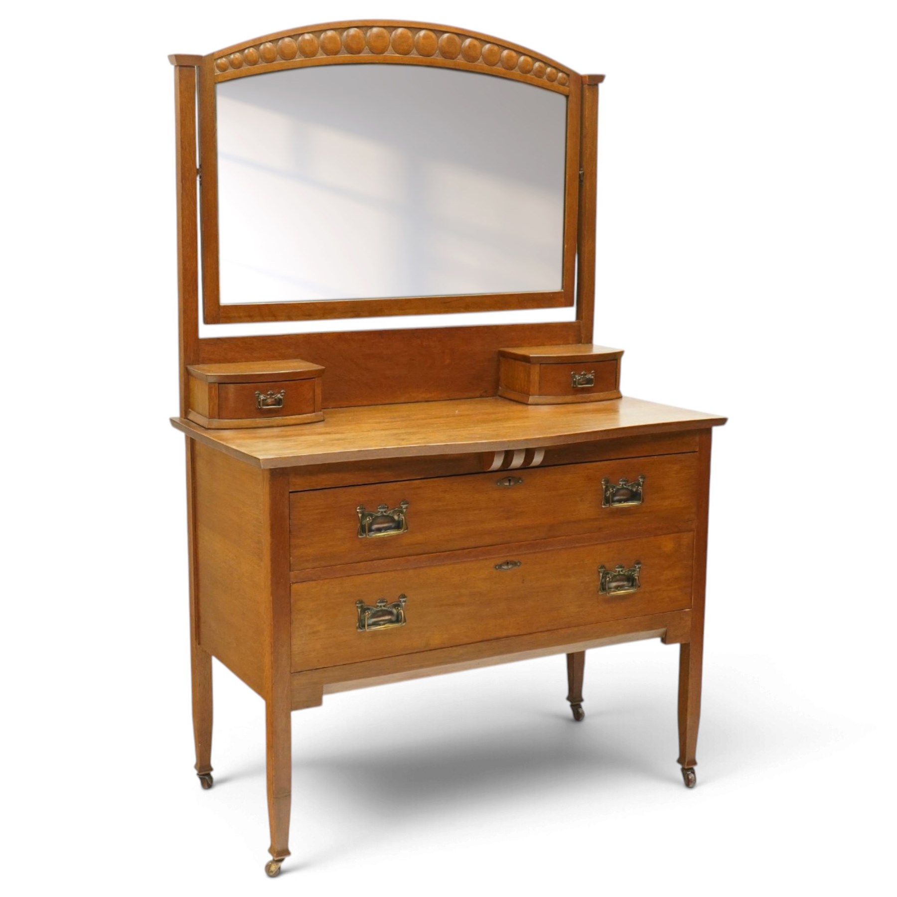 Manner of Liberty & Co. - Arts & Crafts oak dressing chest, the arched swing mirror with roundel-carved frieze and bevelled plate, serpentine-front top with two raised trinket drawers, fitted with two drawers with brass Art-Nouveau poppy handle-plates, on square tapering supports with castors