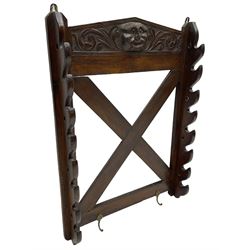 Victorian oak wall hanging whip or stick rack, sloped arch pediment carved with central grotesque mask with extended scrolling foliage, the upright supports cut with rests, x-framed back, two lower hooks