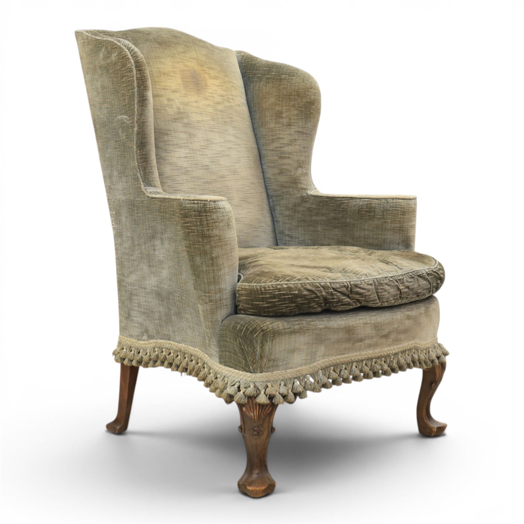 Pair of Georgian style wing armchairs, upholstered in green velvet, each with shaped wing backs, padded scroll arms and loose seat cushions, raised on cabriole supports terminating in pad feet, one with tasselled fringe