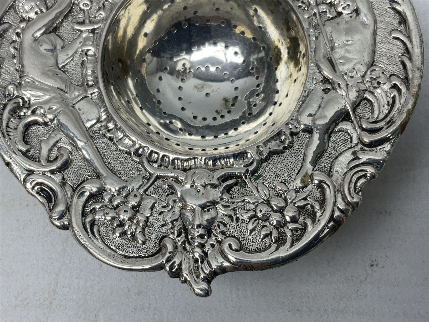 Continental white metal strainer, of shaped form embossed with classical figures, putto, musical trophies and fruiting vines, indistinctly marked, L12.5cm