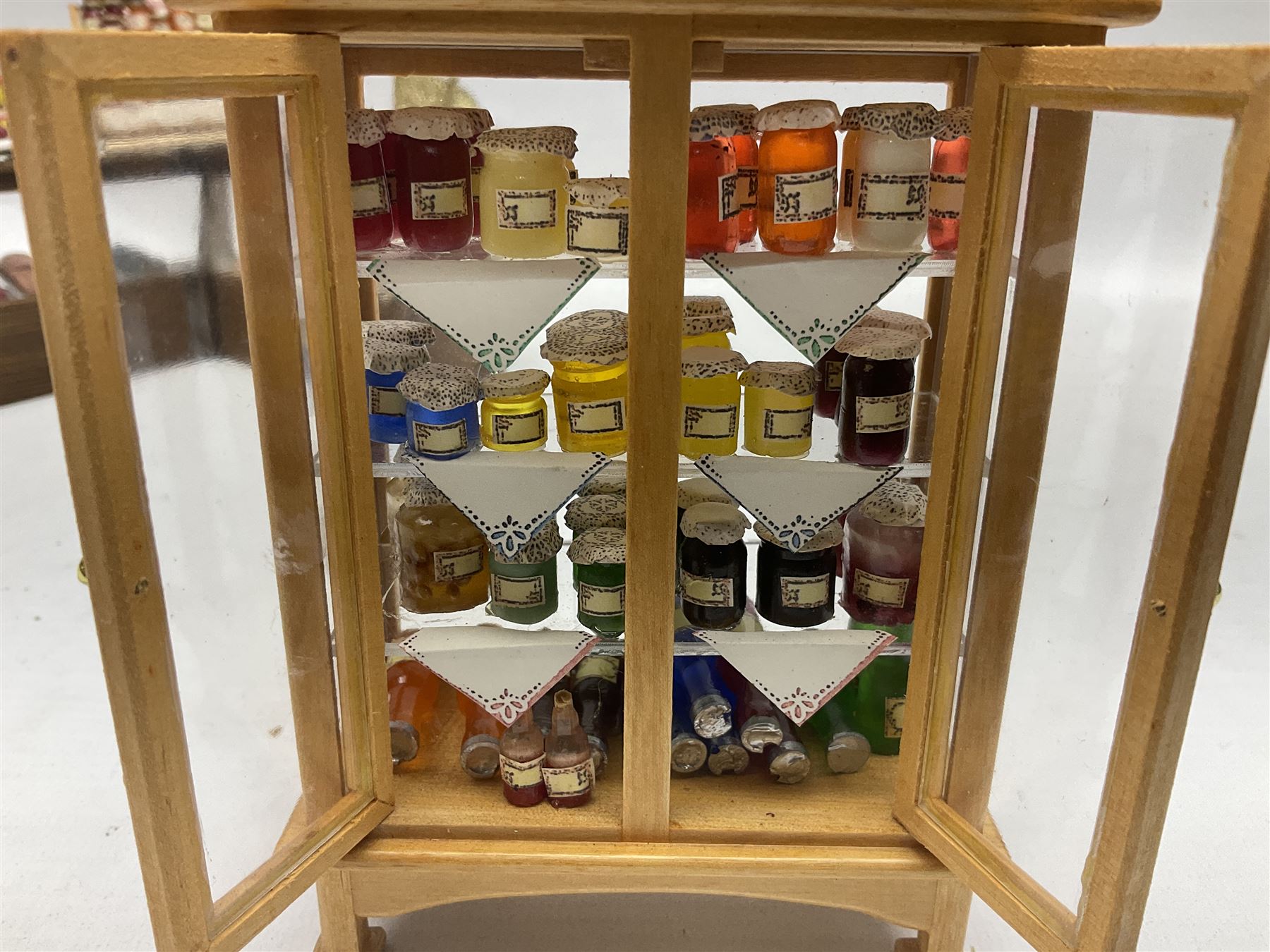 Collection of miniature dolls house grocery shop furniture and produce, comprising glazed corner counter with brass style cash register, shelved preserves unit, pies, cheese board and trays of meats, pair of white cheese counters, biscuit display with six tins, glazed preserves cabinet, pair of shop chairs and four further cheese boards complete with grapes and label flags