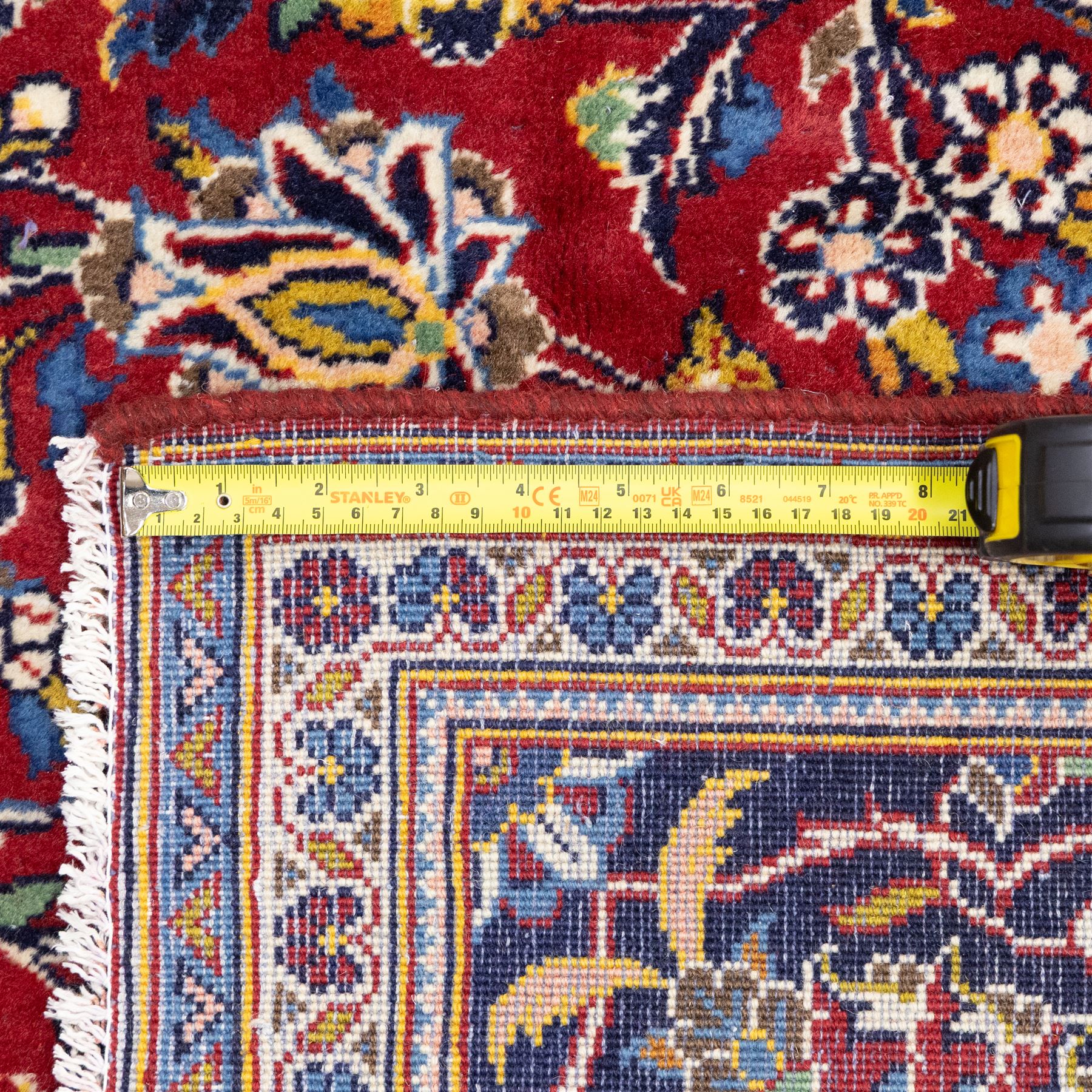 Central Persian Kashan hand-knotted rug, rich red field with a central lobbed medallion surrounded by finely drawn scrolling floral sprays, palmettes, and rosettes, enclosed by a broad dark blue floral border with multiple guard stripes and cotton fringe ends 