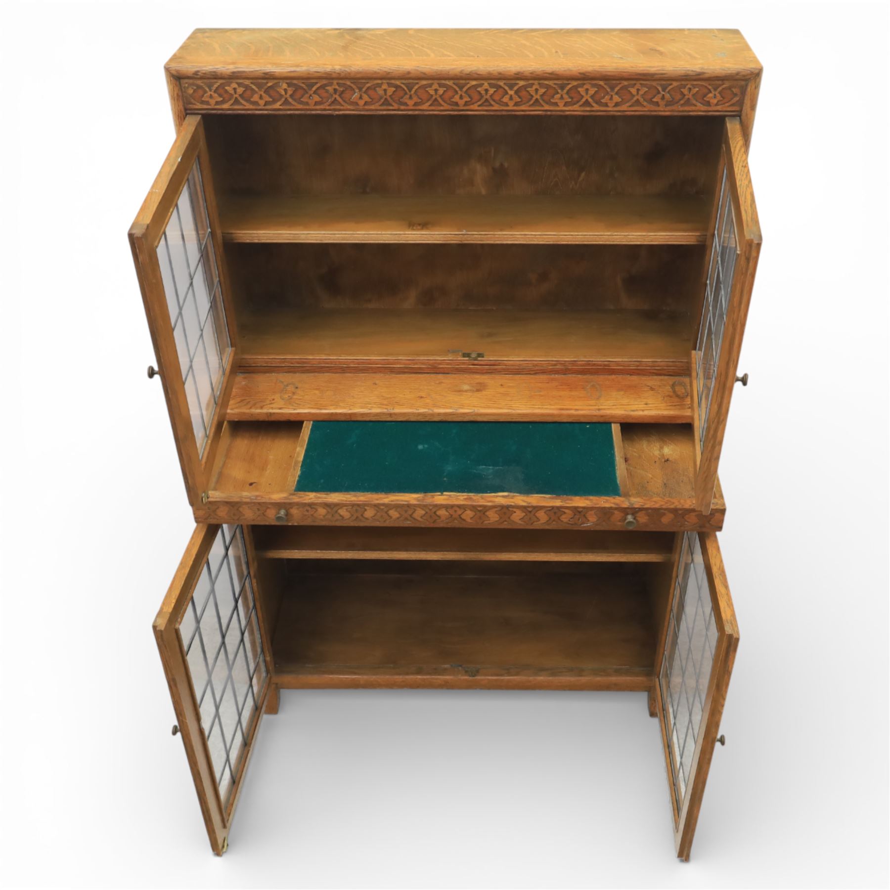 Early 20th-century Arts and Crafts glazed oak bookcase, upper section fitted with a pair of leaded glazed doors enclosing a fixed shelf above a projecting frieze drawer interior with baize-lined writing surface flanked by two compartments, carved with interlaced decoration, lower section fitted with a further pair of leaded glazed doors enclosing a single shelf, raised on short stile feet 