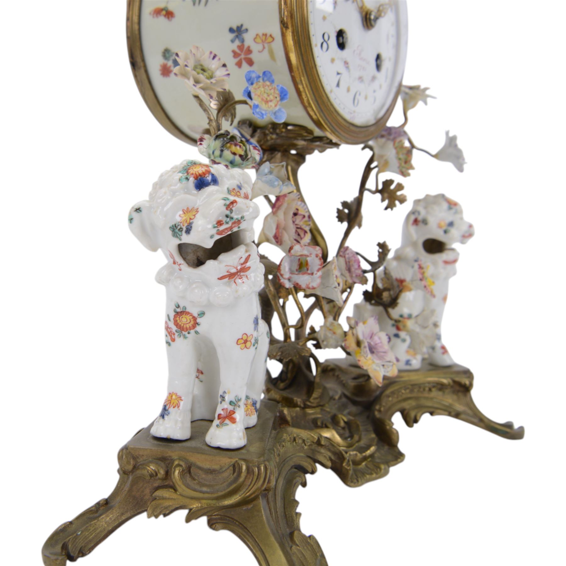 French- early 20th century 8-day Chinoiserie brass and porcelain mantel clock in the 18th century style, brass base with splayed feet profusely decorated with clusters of painted porcelain flowers and two decoratively painted Chinese foo dogs, movement enclosed in a circular porcelain movement case illustrated with birds of paradise and oriental motifs, with a white enamel dial Arabic numerals, minute markers, pierced gilt hands and a floral swag, rack striking Parisian movement striking the hours and half-hours on a bell. With a visible sunburst pendulum and key.