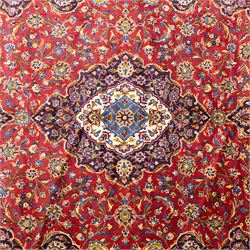 Central Persian Kashan hand-knotted carpet, crimson central field with lobbed medallion in navy blue and ivory ground, surrounded by a dense all-over floral design of scrolling vines, palmettes, and stylized blossoms, enclosed within a deep navy main border, flanked by multiple guard stripes with fringe ends 


