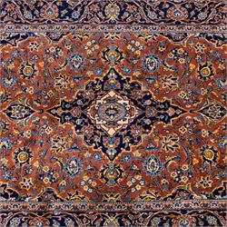 Central Persian Kashan hand-knotted rug,  terracotta field with central shaped ivory and dark blue medallion with palmettes, floral sprays and scrolling vine ornament, with conforming corner spandrels enclosed by a broad navy border of palmettes and flowering vine between multiple guard stripes, with cotton fringe ends 