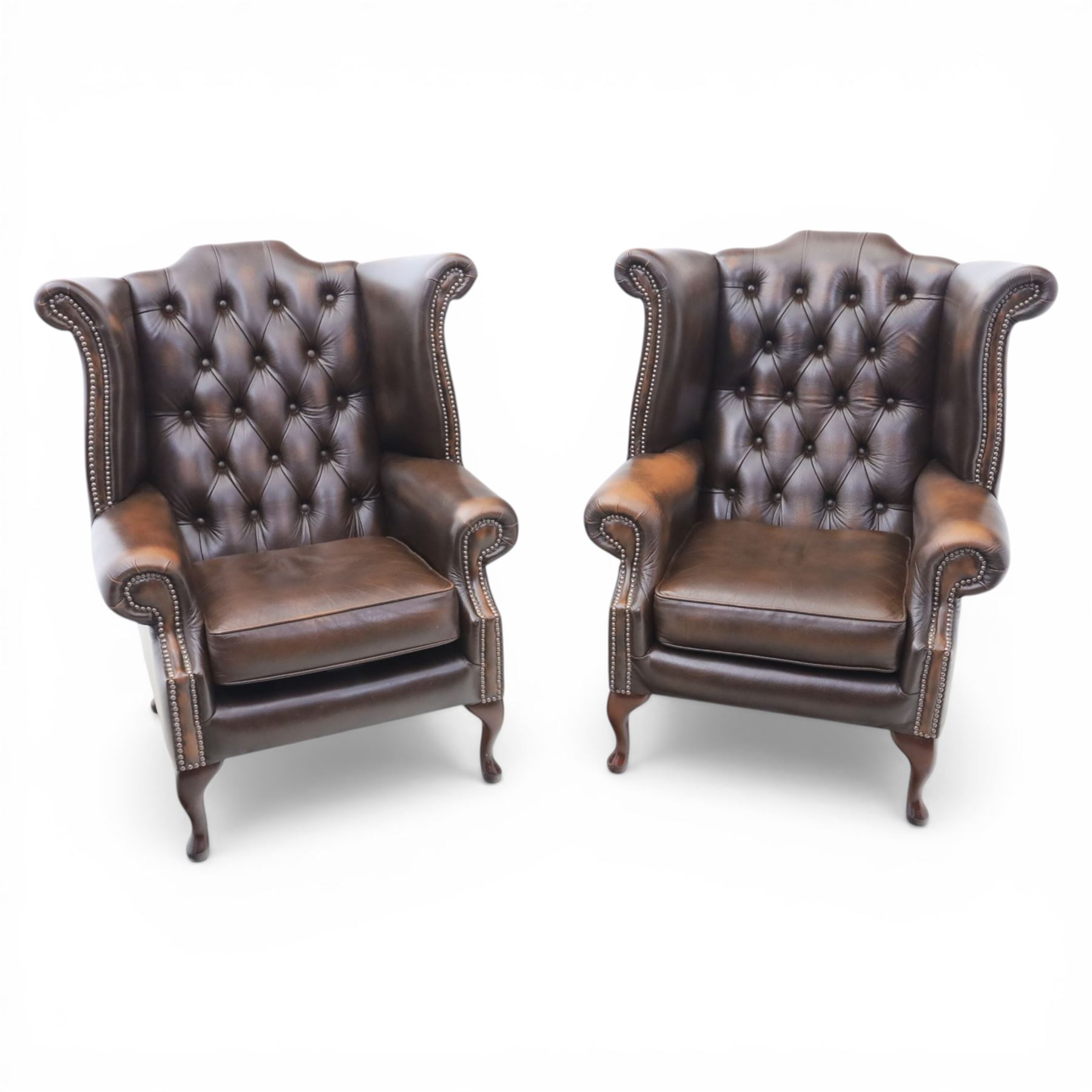 20th century Chesterfield style brown leather wingback armchairs, with button upholstered backs and wings, shaped scroll arms with studded borders, loose seat cushions, raised on cabriole supports with pad feet