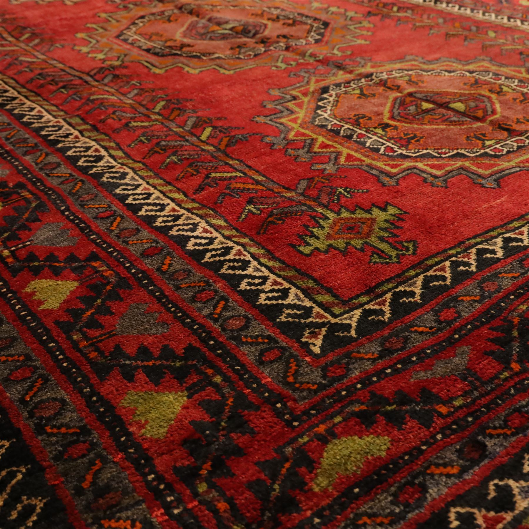 North East Persian Kurdish hand-knotted carpet, madder red field with three vertical and central stepped octagonal medallions enclosing angular geometric motifs with scattered tribal devices, framed by multiple narrow borders with latch-hook patterns, enclosing a bold main border of repeating geometric motifs, palette with indigo, dark brown and soft green tones, 