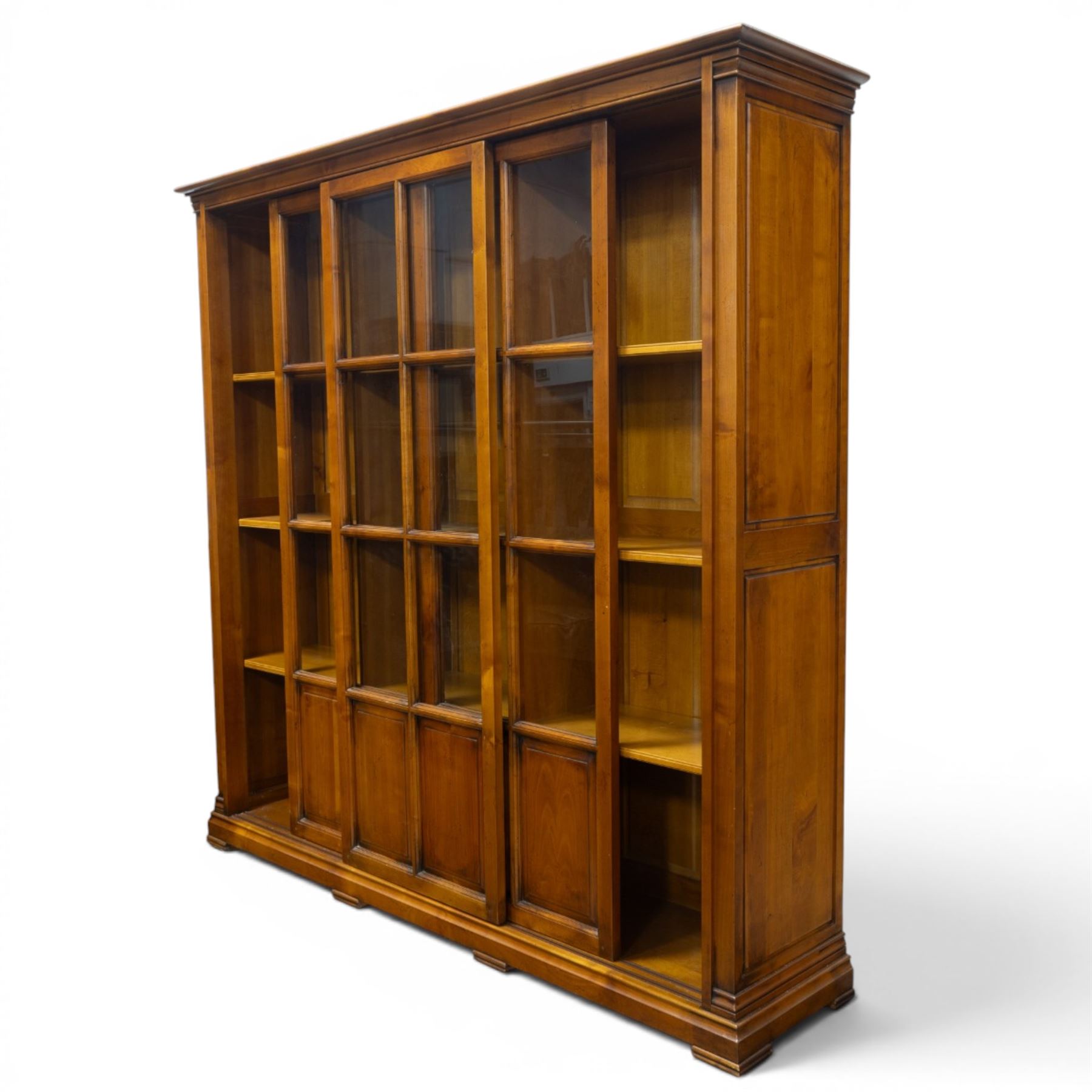 Wesley Barrel - large cherry wood bookcase, with projecting cornice and fielded panelled sides, with three sections comprising three fixed shelves, sliding glazed and panelled doors, moulded plinth base
