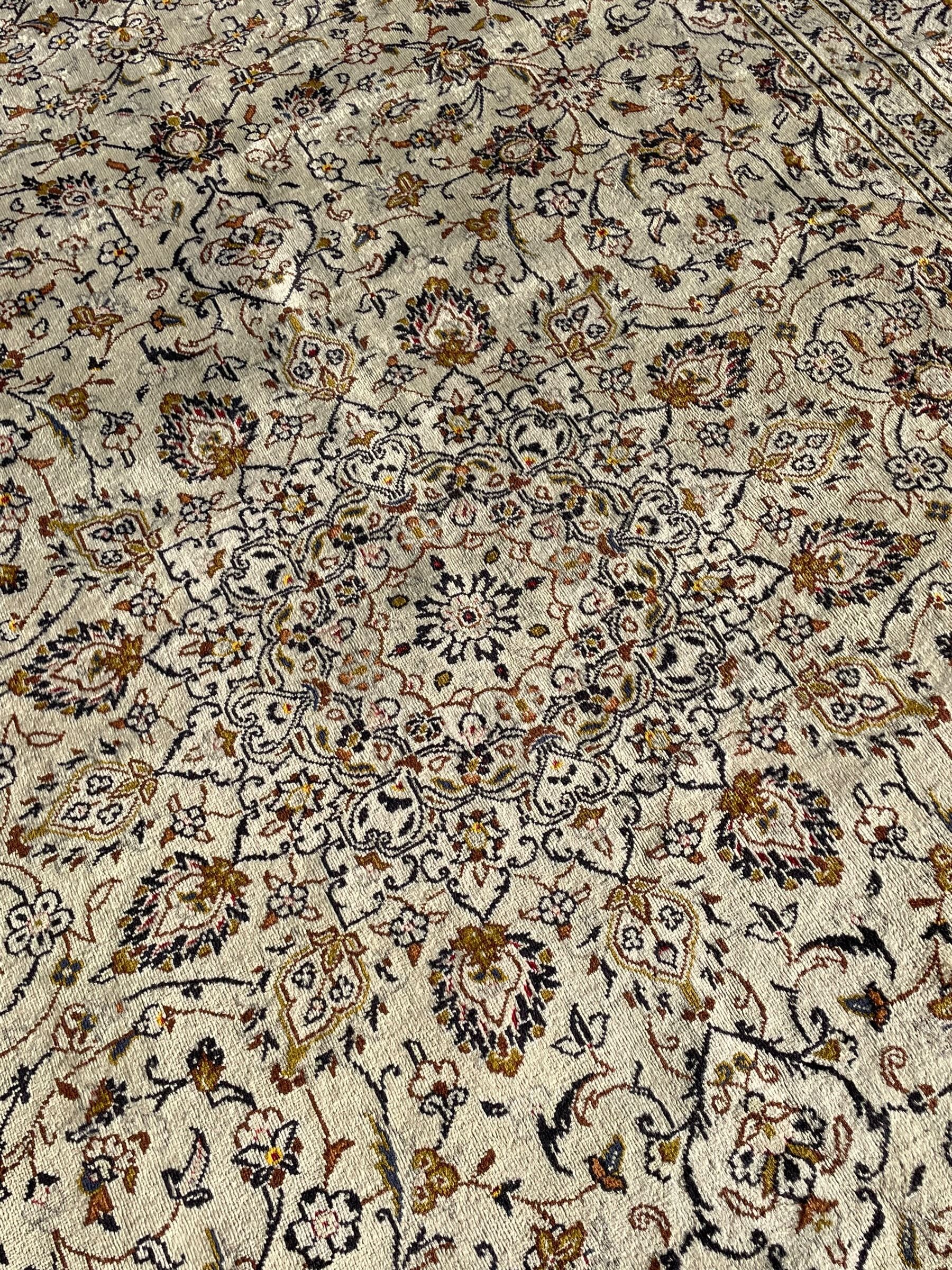 Persian Kashan ivory ground carpet, central medallion decorated with sixteen arranged palmette motifs, within a field decorated with swirling leafy branches and stylised plant motifs, the main border decorated with matching swirling pattern, within floral pattern guard stripes 
