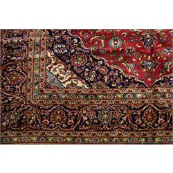 Persian Kashan hand-knotted carpet, madder red ground centred by shaped dark navy and ivory floral medallion with pendants, surrounded by scrolling vines, palmettes and flowerheads, dark navy spandrels, enclosed by dark navy floral border with multiple narrow floral guard stripes