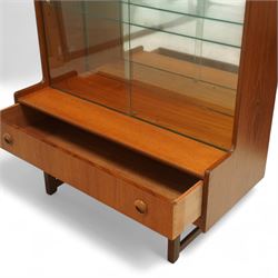 Turnidge of London mid-century teak display cabinet, rectangular top with a pair of glass sliding doors enclosing two adjustable shelves with mirrored back, projecting lower section incorporating one full-width drawer with recessed circular handles, raised on square-section legs with lateral stretchers  