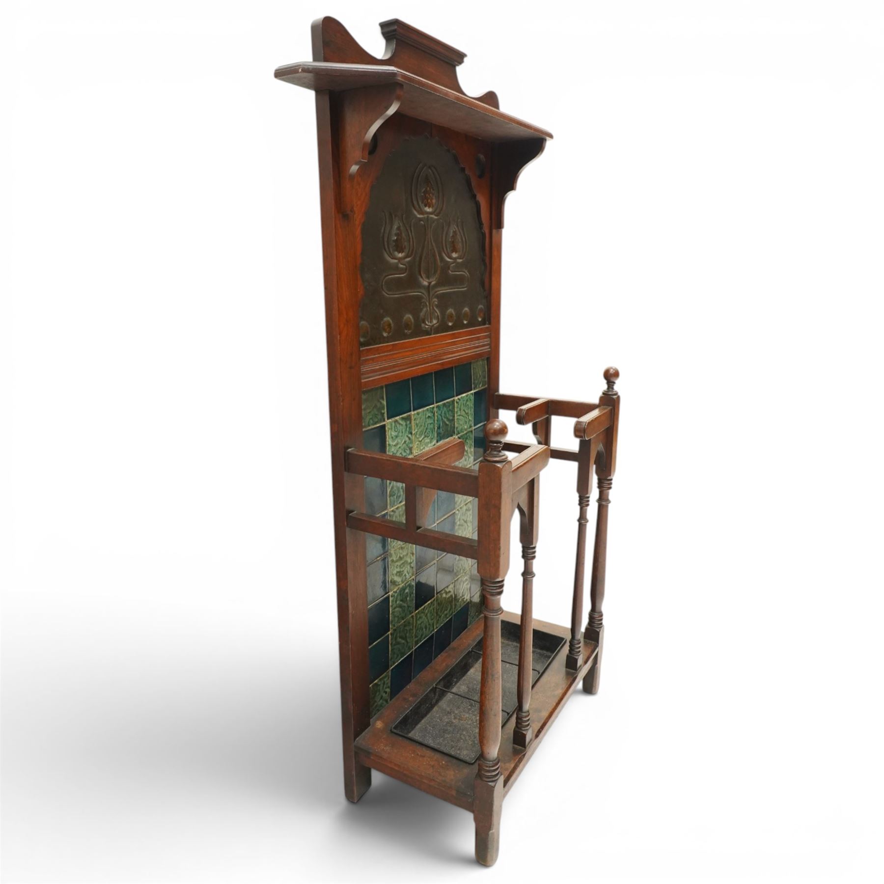 Shapland & Petter of Barnstaple - Arts & Crafts mahogany tile-backed umbrella or stick hall-stand, model no.114, the shaped back with single shelf over an Art Nouveau embossed patinated copper panel and turquoise tiled back, with turned supports and dividers over inset metal drip tray

Notes: A comparable Shapland & Petter stick stand of this model is recorded in Daryl Bennett, Shapland & Petter Ltd of Barnstaple, Museum of Barnstaple and North Devon, p.84, cat. no.5.37, and the model is also listed in Bennett, Liberty’s Furniture 1875-1915, p.298, cat. no.8.6. Examples of this model are known to have been retailed by Liberty & Co.