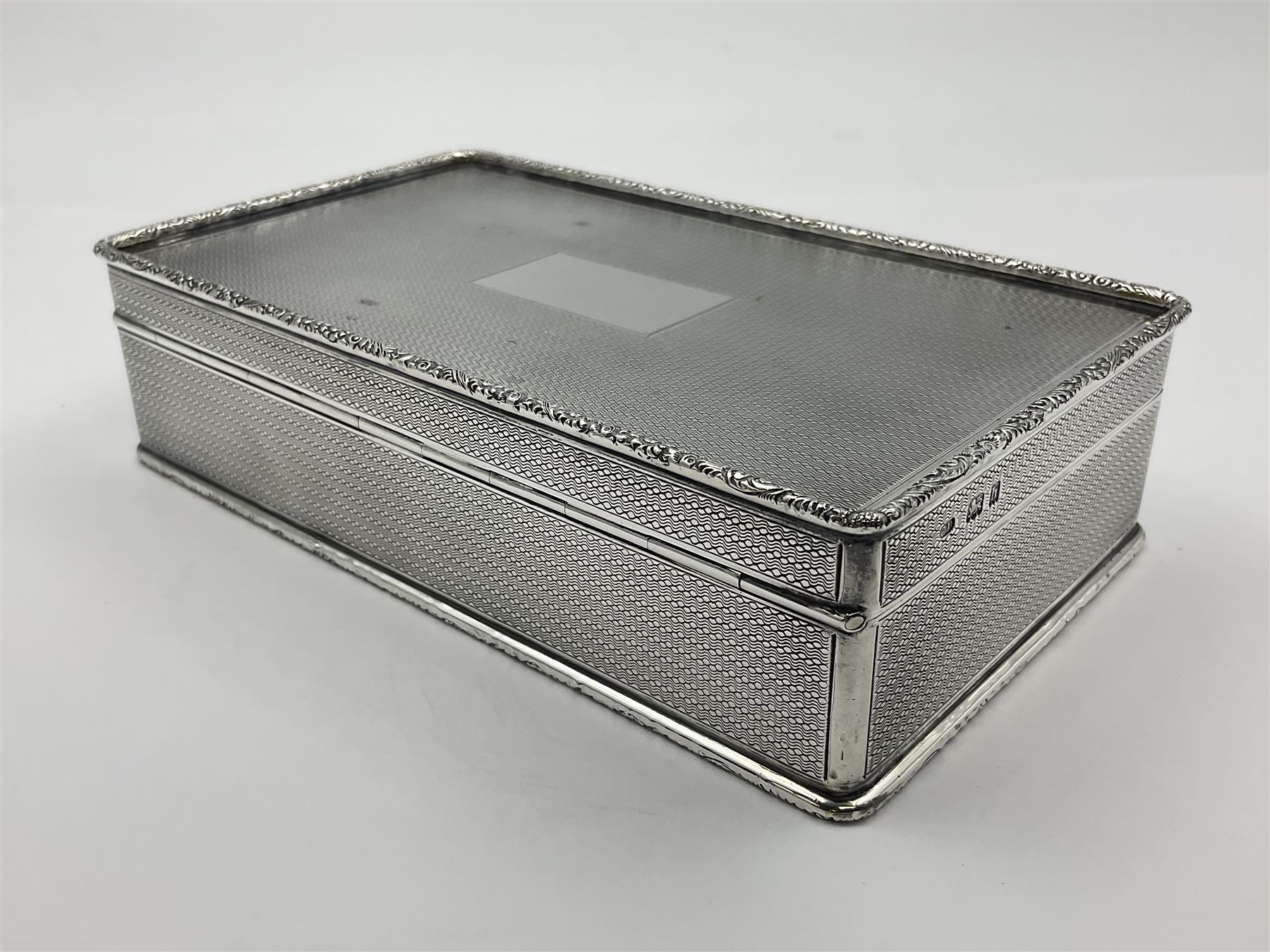1920's silver table cigarette box, of rectangular form with engine turned decoration to the exterior, the hinged cover with central vacant rectangular panel and raised foliate rim, opening to reveal a gilt interior with later plush lining, hallmarked Charles Henry Dumenil, London 1923, H3.5cm L14.5cm D8.5cm