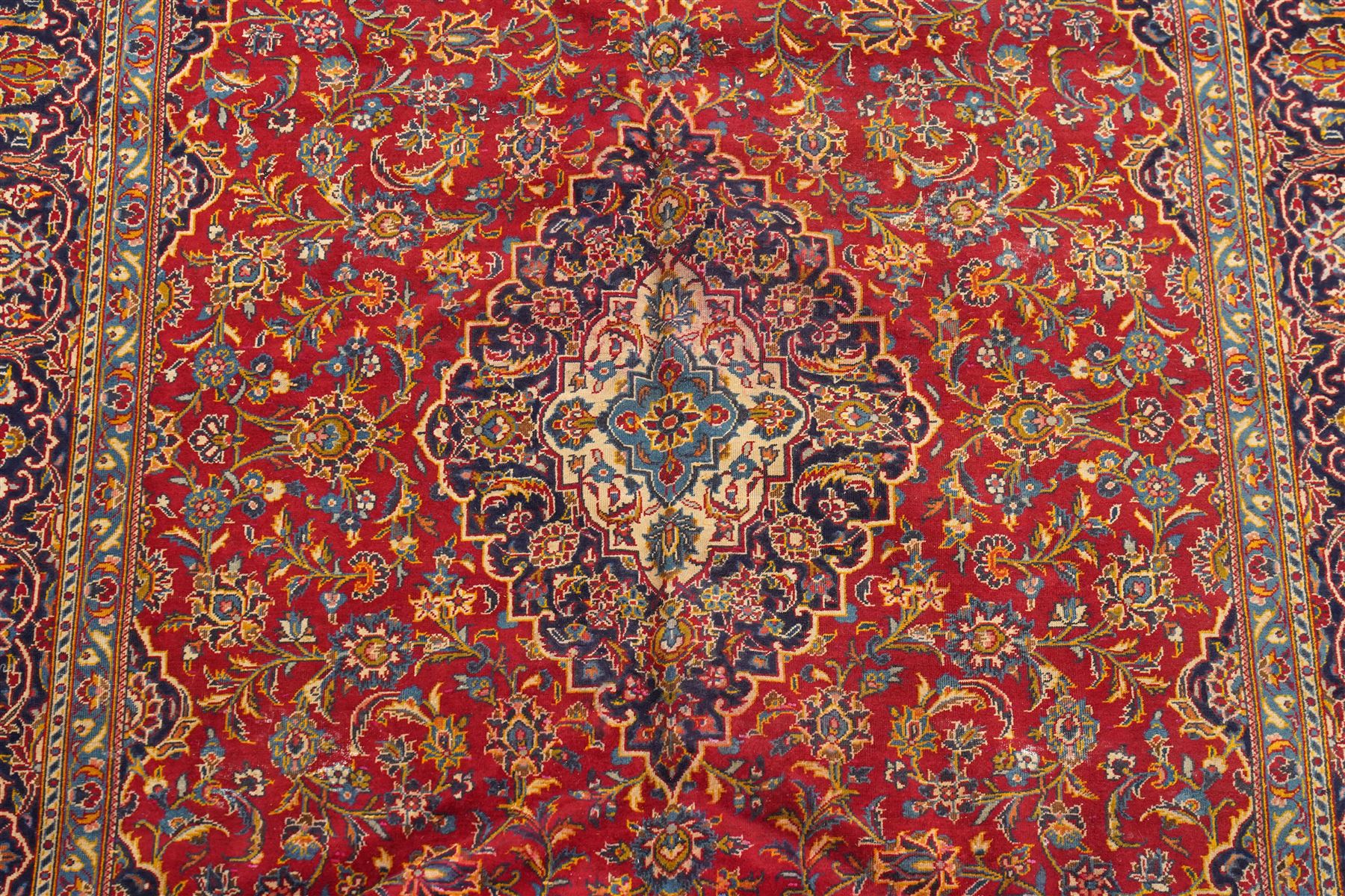 Persian Kashan hand-knotted carpet, madder red ground centred by shaped dark navy, ivory and pale blue floral medallion with pendants, surrounded by scrolling vines, palmettes and flowerheads, dark navy spandrels, enclosed by dark navy floral border with multiple floral guard stripes