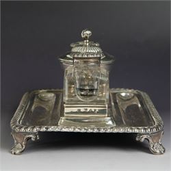 George IV silver desk stand, of rectangular form with rounded corners and oblique gadrooned rim, upon four embossed pad feet, with central pedestal dish of part fluted form with domed lid engraved with crest and with bud finial, with two recesses either side housing faceted glass inkwells, each with silver collars, hallmarked John & Thomas Settle, Sheffield 1825 & 1827, H11cm