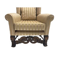 William and Mary style wingback armchair, upholstered in gold striped fabric, on walnut scroll carved supports joined by scroll and foliate carved middle rail, turned H-shaped stretchers, on castors