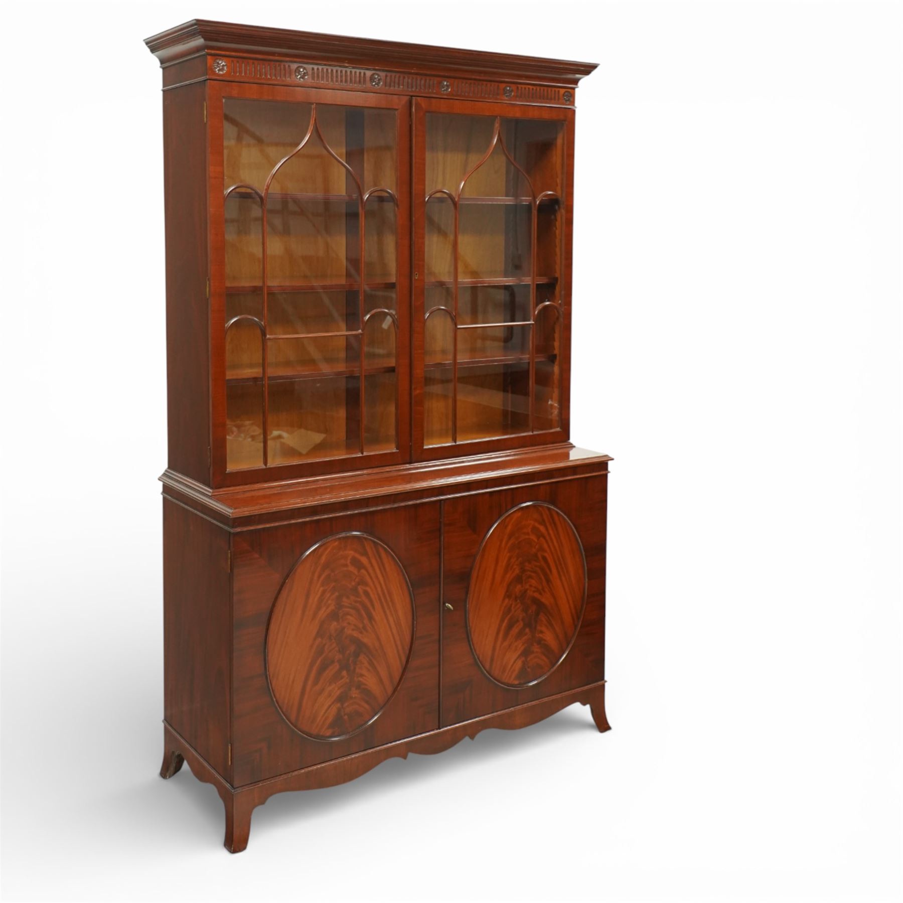 William Tillman - Early 20th century mahogany display cabinet, moulded cornice above a frieze carved with roundels and fluting, pair of astragal glazed doors enclosing adjustable shelving, the lower section fitted with twin panelled cupboard doors centred with oval veneered panels, shaped apron and bracket feet