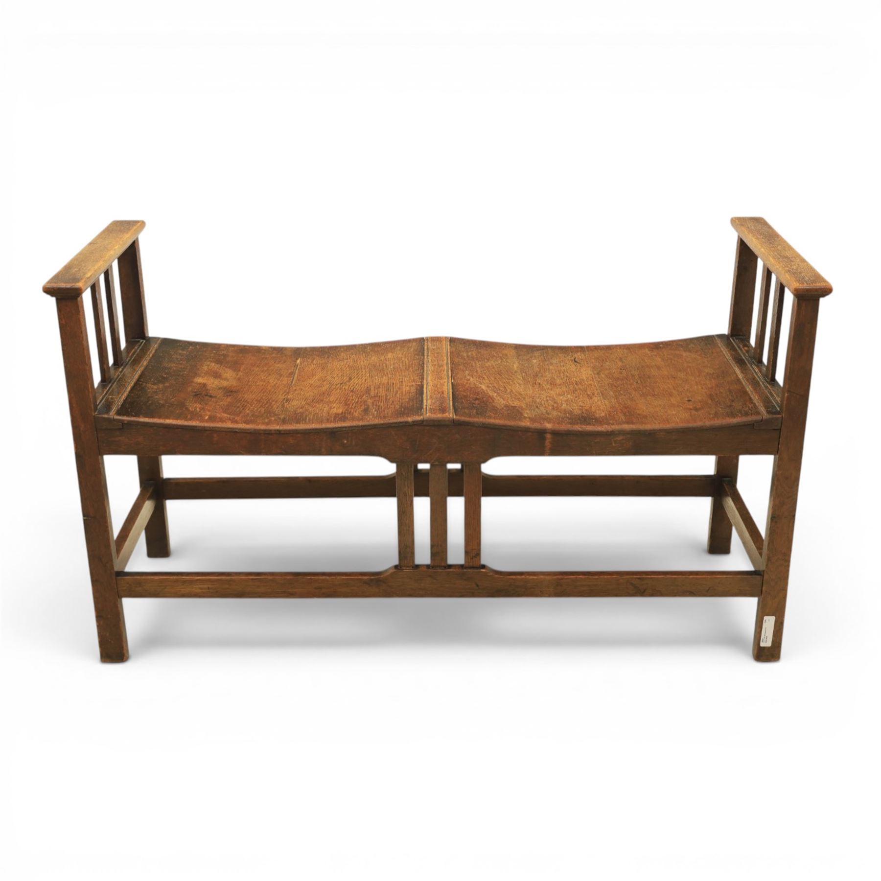 Arts and Crafts oak window bench, with flat armrests flanking two dished seats, raised on square supports united by stretchers