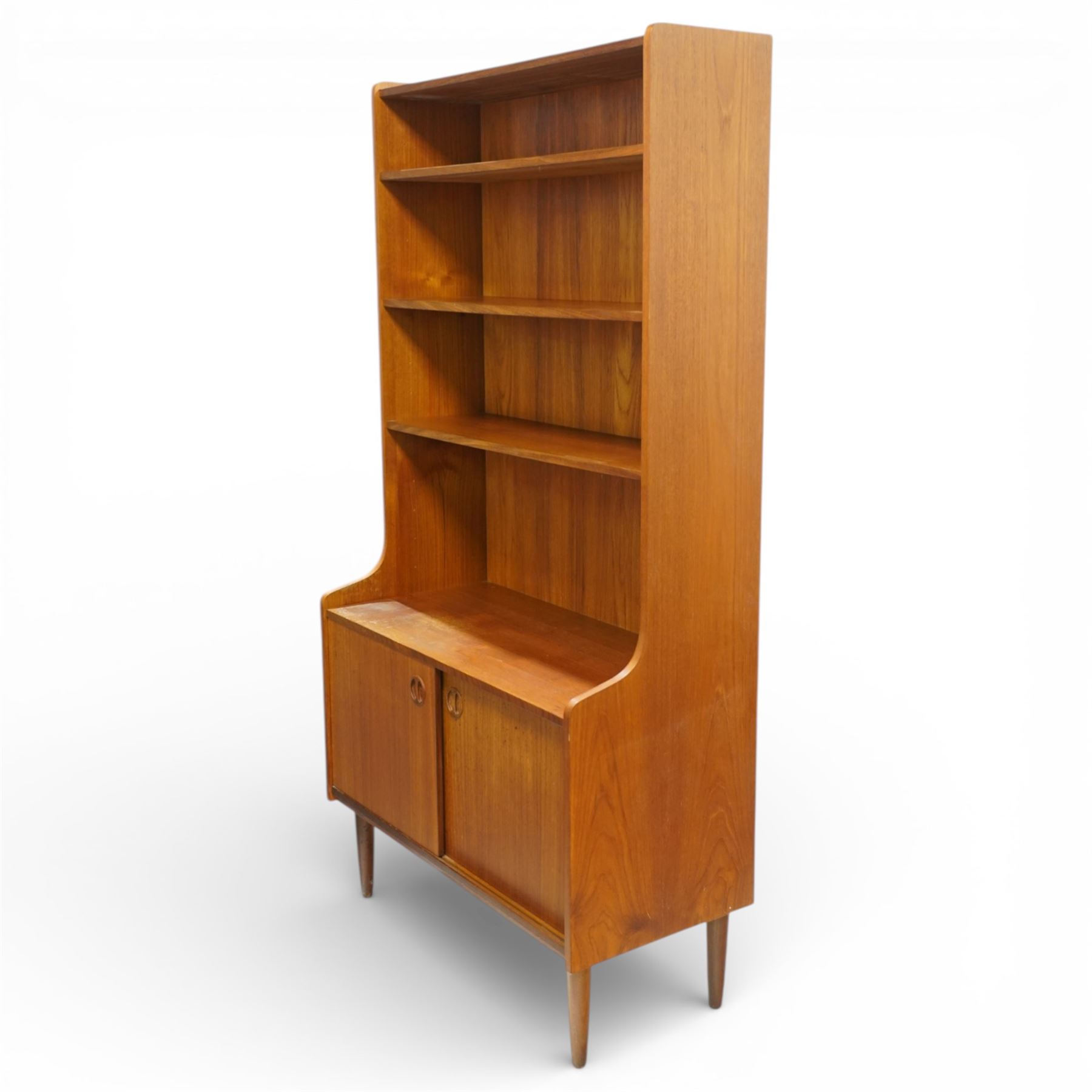 Mid 20th century Scandinavian teak bookcase cabinet in the manner of Johannes Sorth, the upper section with three open shelves over a shaped fall to a lower cupboard enclosed by sliding doors with recessed circular handles, raised on tapering supports