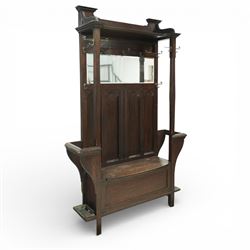 Arts and Crafts design oak settle, with projecting moulded cornice over a boarded back fitted with a rectangular bevelled mirror plate and a row of wrought iron coat hooks, the panelled back with arched fielded panels, flanked by shaped umbrella or stick stands with removable drip trays, the plank hinged seat over a panelled box base, on square section supports