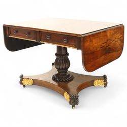 Regency rosewood sofa table, rectangular top with variegated rosewood veneers, hinged D-shaped leaves, over a set of dummy drawers to the back and full-size drawers to the front, with turned knob handles and gilt beaded moulding on frieze above elegant turned, tapering, and fluted, pedestal with a well-carved collar, supported on a large, tripartite concave platform base terminating in richly carved lion paw feet with gilded acanthus leaf carving, on concealed brass castors, circa 1815, length extended 161cm 

