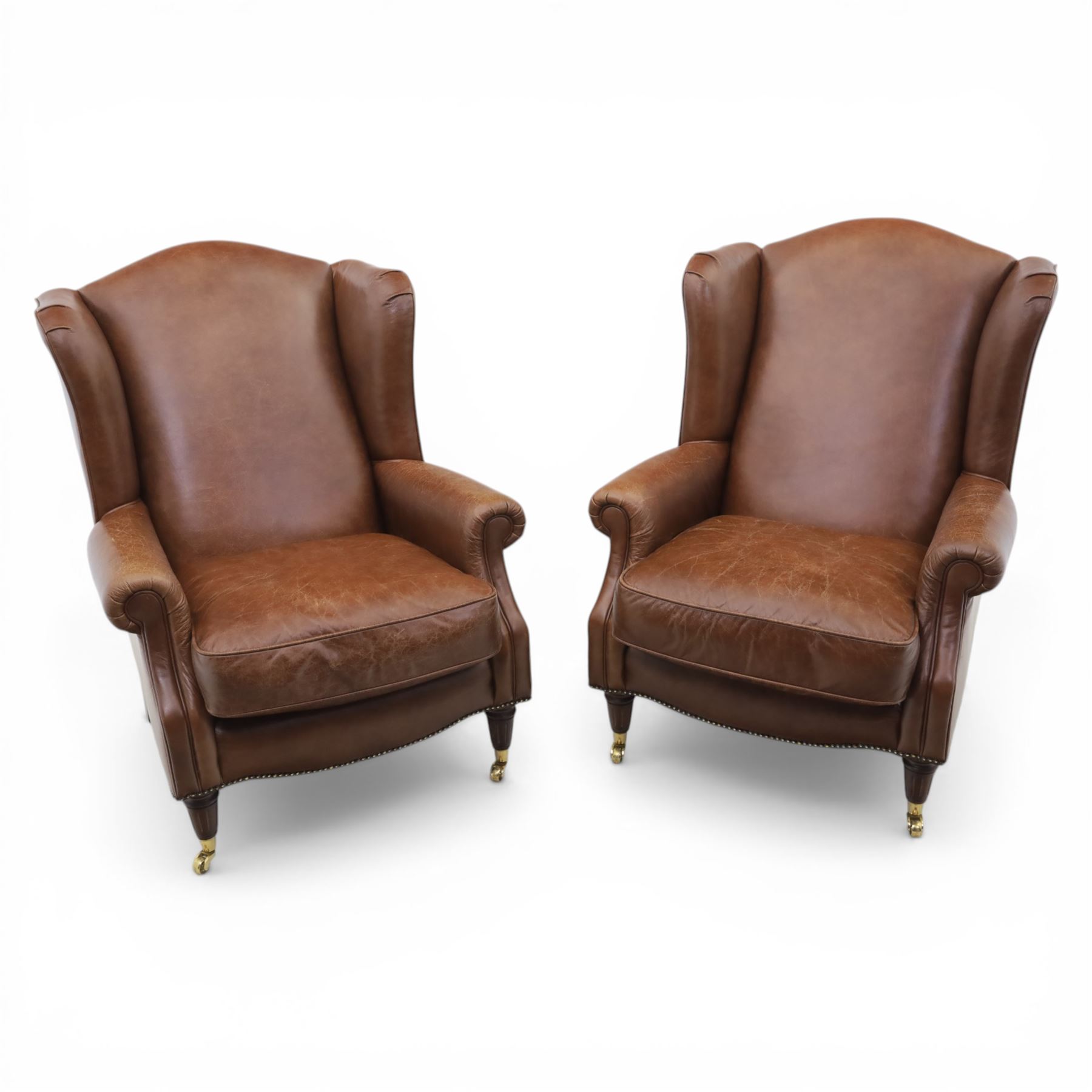 Laura Ashley - Pair of brown leather wingback armchairs, with scrolled arms and loose cushion seat, brass studded trim, on cylindrical tapering legs terminating in castors