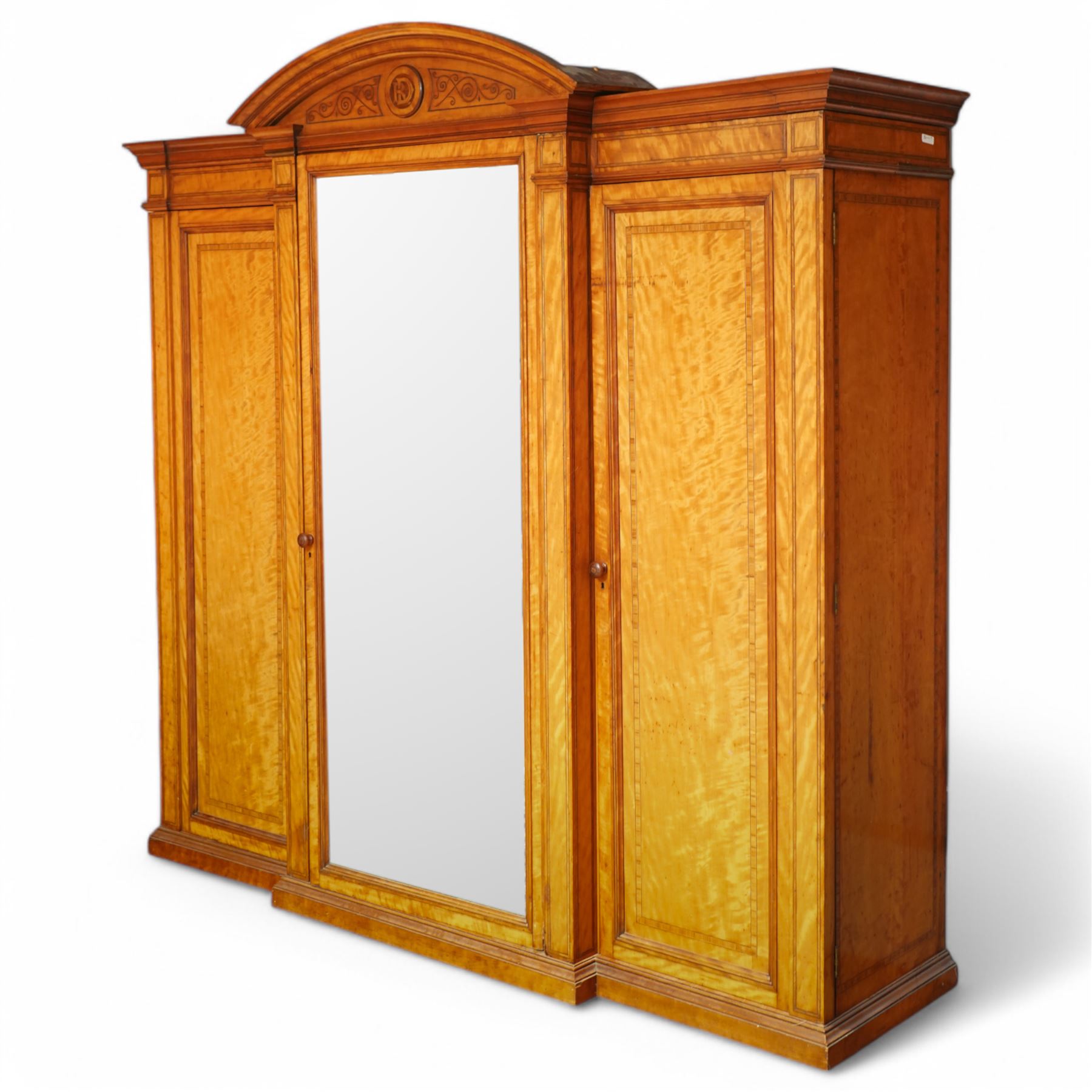 Victorian neoclassical style satinwood triple-section wardrobe, the central mirror section and arched pediment with carved monogram flanked by two panelled crossbanded doors, revealing two drawers and grooves for shelves, the hanging cupboards each with a single drawer, the whole supported on a plinth base