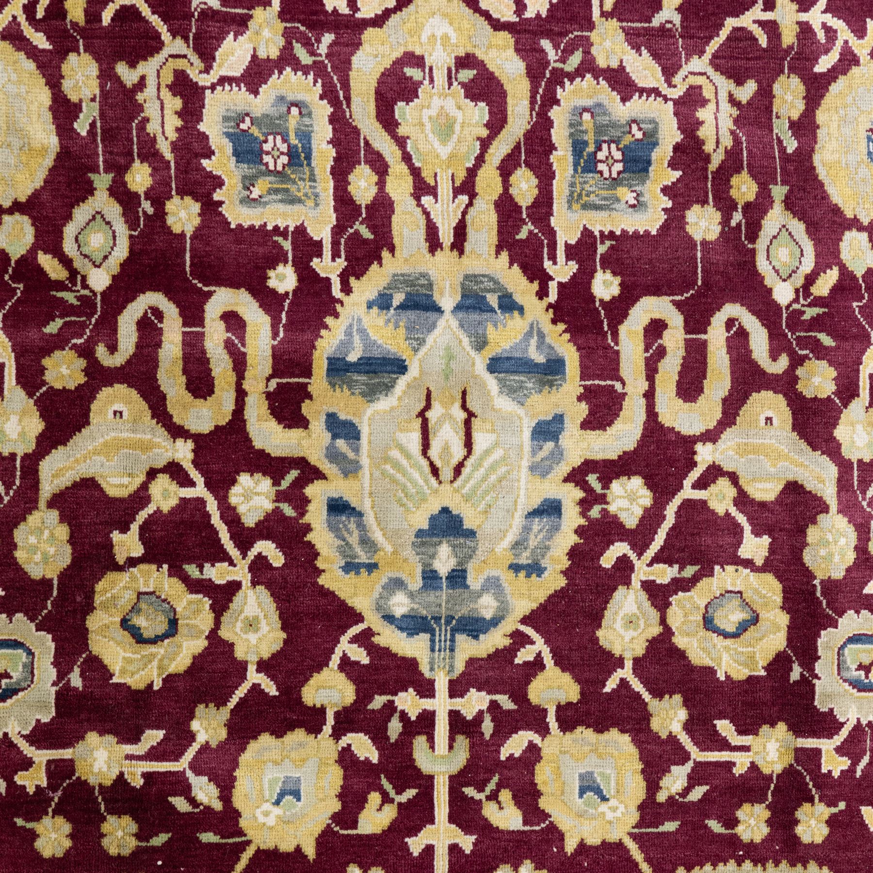 Agra design wool carpet, burgundy field decorated with large scrolling floral vines, palmettes and flowerheads, enclosed by blue main border with oversized stylised palmettes and narrow guard stripes
