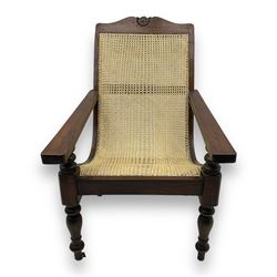 Late 19th century caned plantation chair, shaped top rail with carved central motif over caned back and seat, extending armrests, on turned front supports