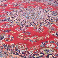 North East Persian Meshed hand-knotted rug, madder red field with central multi-lobbed medallion in shades of indigo, ivory, and sofa pastel tones surrounded by scrolling floral vines, palmettes, stylized blossoms, floral sprays, and cartouches, framed by Indigo spandrels, enclosed within a broad dark blue main border, flanked by multiple narrow guard stripes, with fringe ends 