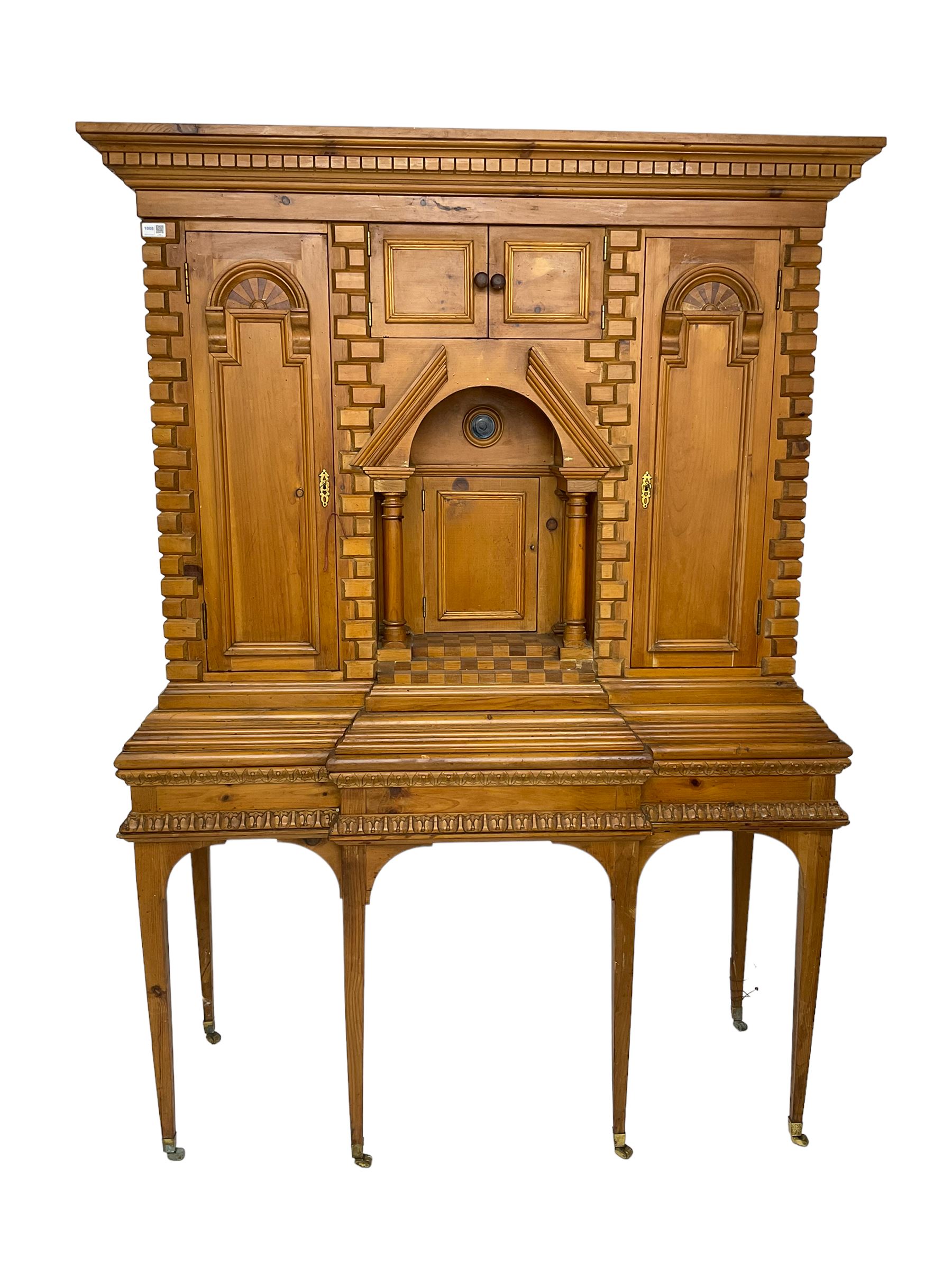 20th century pine architectural collector's cabinet on stand, projecting dentil cornice over various compartments, central recessed arch with turned pillar supports and sloped arch pediment, fitted with four cupboards, false brickwork uprights to front and corners, central hidden drawer revealing cube work surface, lower moulded plinth, the stand with foliate carved mouldings, on six square tapering supports with brass castors
