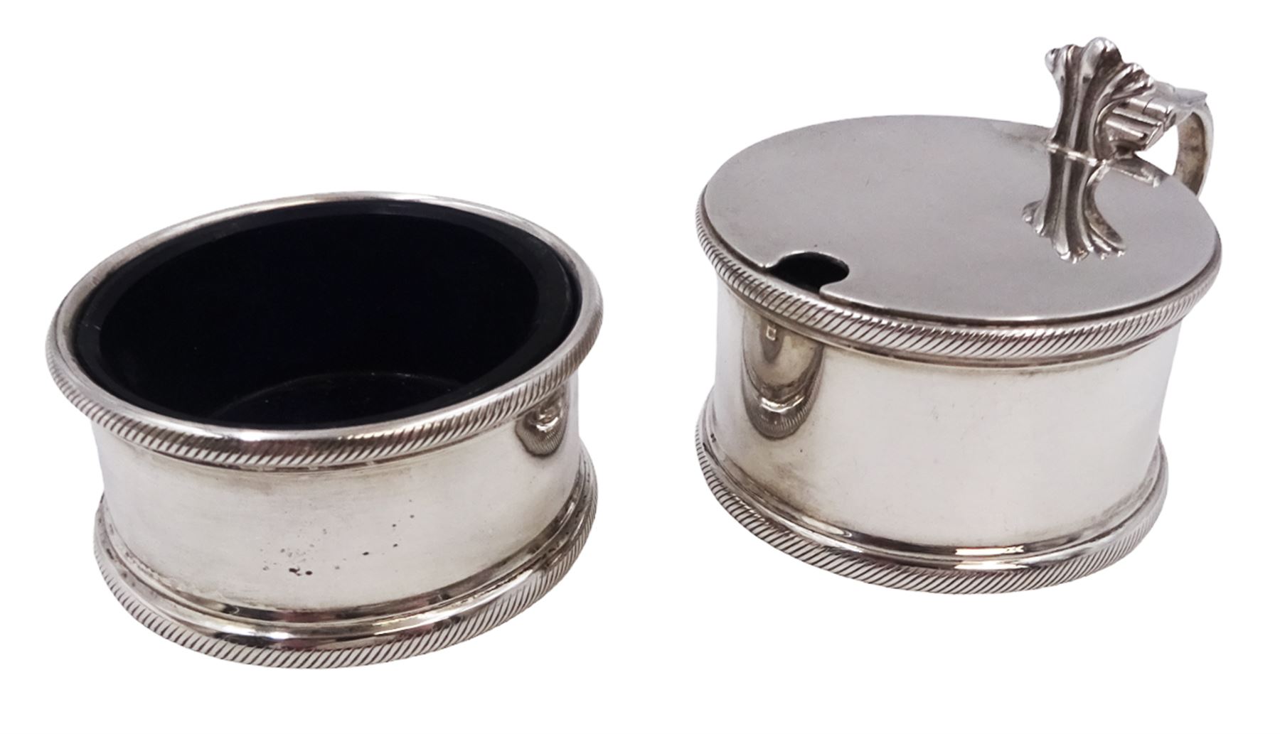 1930s silver part cruet set, comprising mustard pot and cover and open salt, both of circular drum form with oblique gadrooned rim, the mustard pot with capped scroll handle and palmette thumbpiece, both with blue glass liners, hallmarked London 1935, maker's mark worn and indistinct