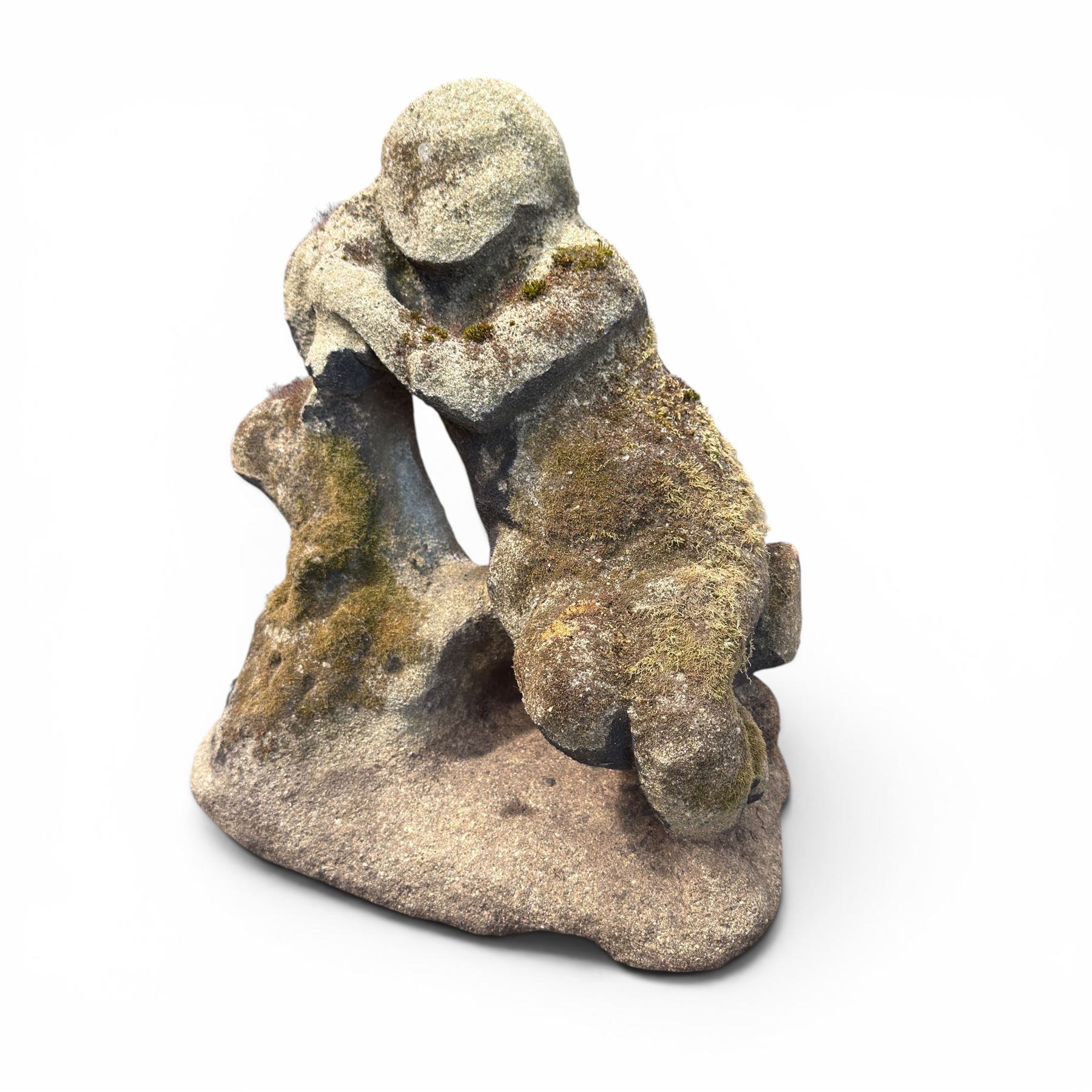 Carved composite stone garden figure, depicting a seated child with folded arms, weathered finish with moss and lichen growth, raised on an integral naturalistic base