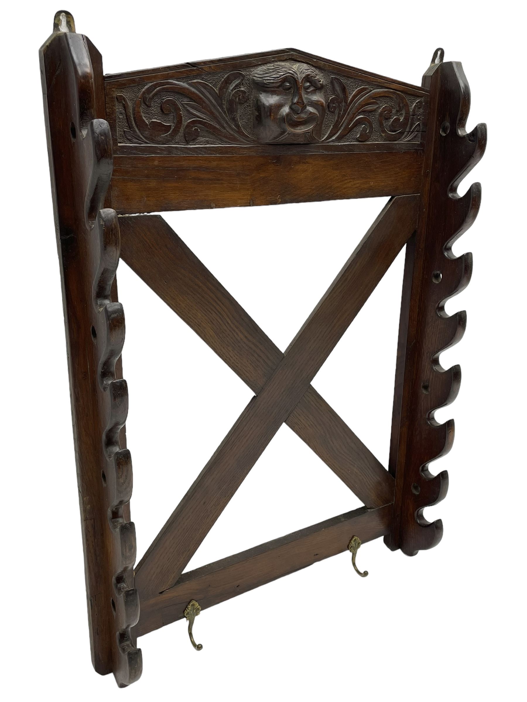 Victorian oak wall hanging whip or stick rack, sloped arch pediment carved with central grotesque mask with extended scrolling foliage, the upright supports cut with rests, x-framed back, two lower hooks
