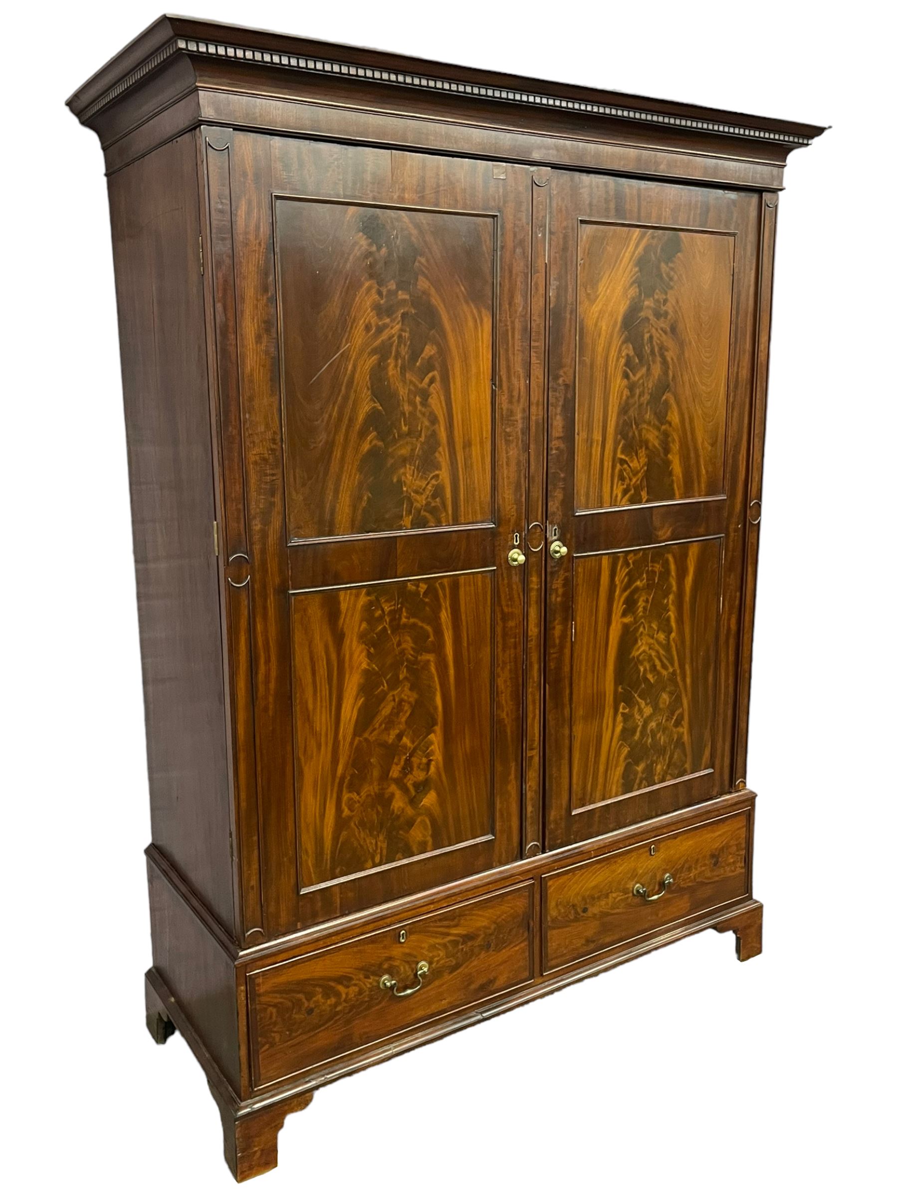 George III mahogany double wardrobe, the projecting cornice with dentil moulding over two panelled doors with mirror-lined interiors, enclosing hanging rails and brass hooks, the base fitted with two drawers, raised on bracket feet