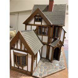 Gerry Welch for Manorcraft Tudor style wooden doll's house, of three-storey multi gabled form with mock beam and plaster walls and simulated leaded glass windows under a felt tiled roof, boarded floors and stairs to each room, curtains to most windows, front entrance door, rear door and single storey extension for kitchen. Furniture includes fitted floral bathroom suite to top floor, well stocked Welsh dresser with copperware etc, tapestry style wall hangings, knole settee, tin bath, metal range, fire-surround, wooden three-piece suite etc; on shaped cobbled base W91cm D61cm H91cm