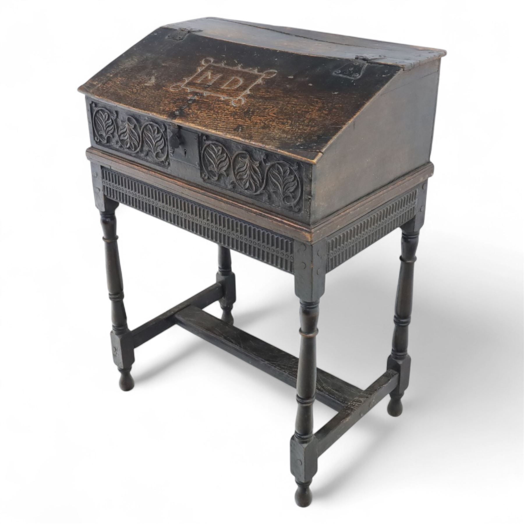 18th century carved oak Bible box on stand, the sloping lid with incised initials ‘MD’ and iron strap hinges, the front carved with stylised leaf and scroll panels enclosing central iron lockplate, on stand with gadrooned frieze, turned legs and plain stretchers