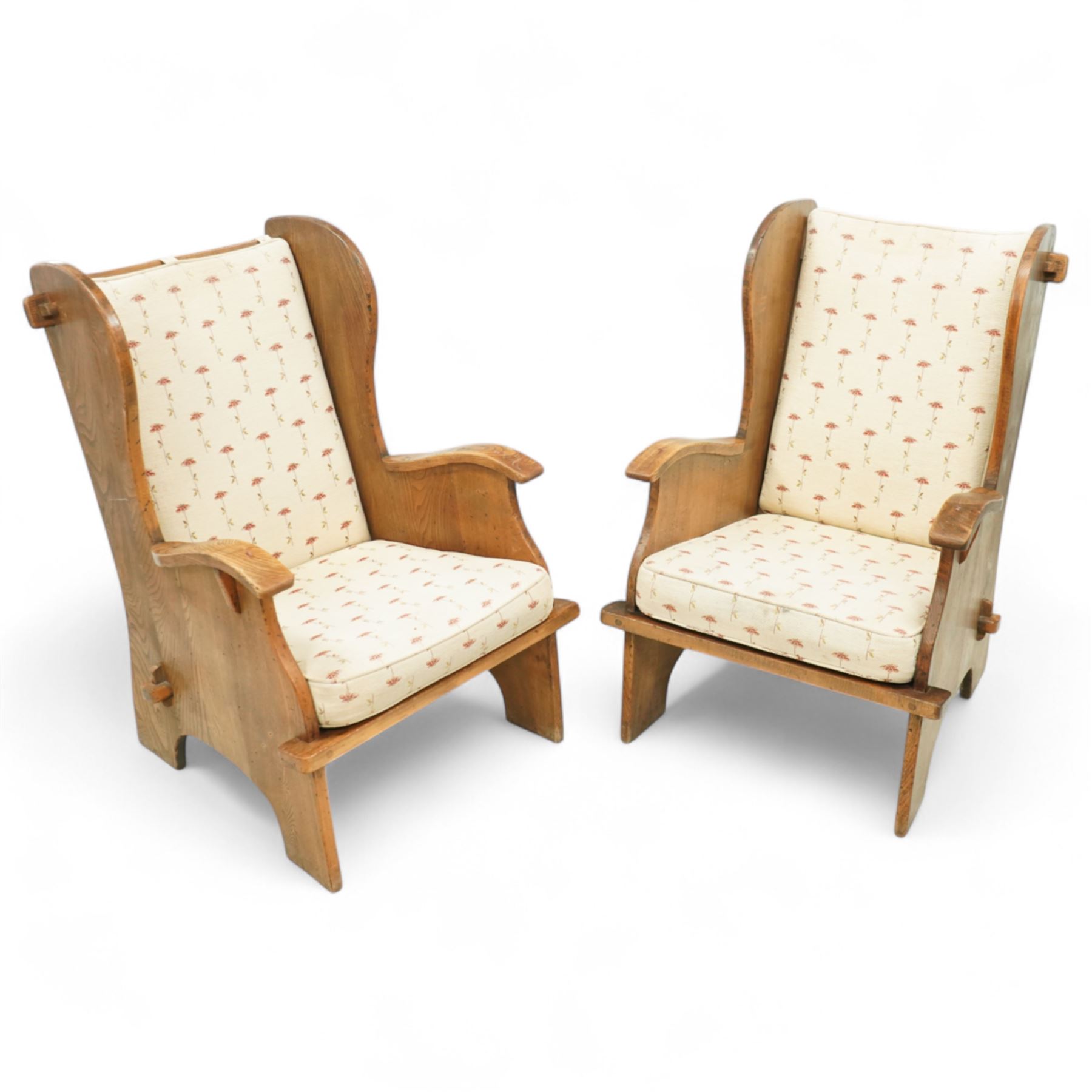 20th century pair of country lambing chairs in light oak, each with high winged back and shaped arms, pegged construction with panelled sides and arched aprons, loose seat and back cushions upholstered in cream floral fabric